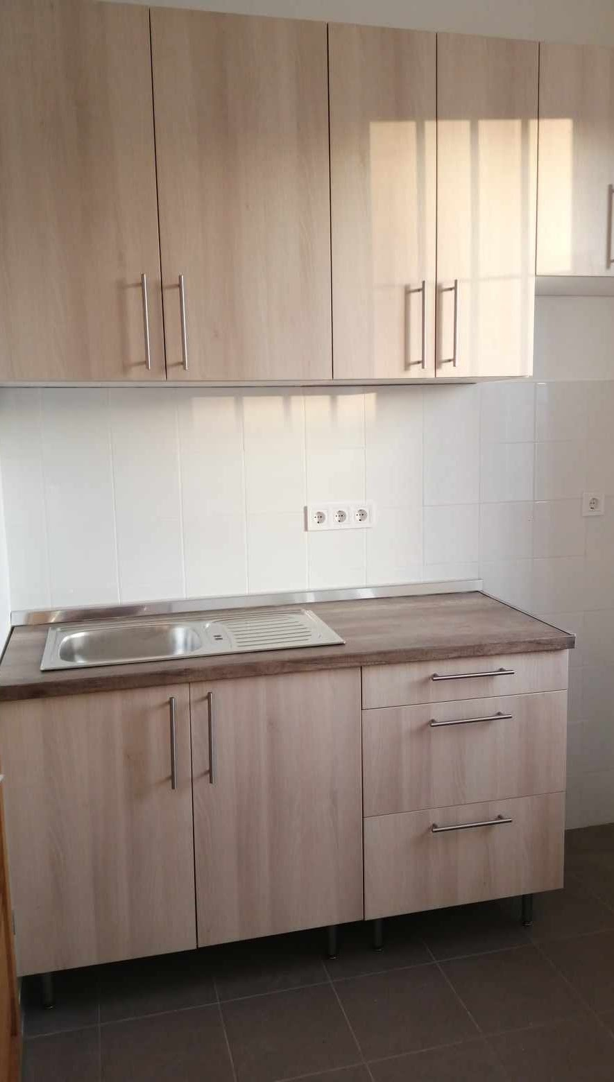 A kitchen with beige cabinets, a wooden countertop, a stainless steel sink with a built-in draining area, white tiled backsplash, and brown floor tiles.