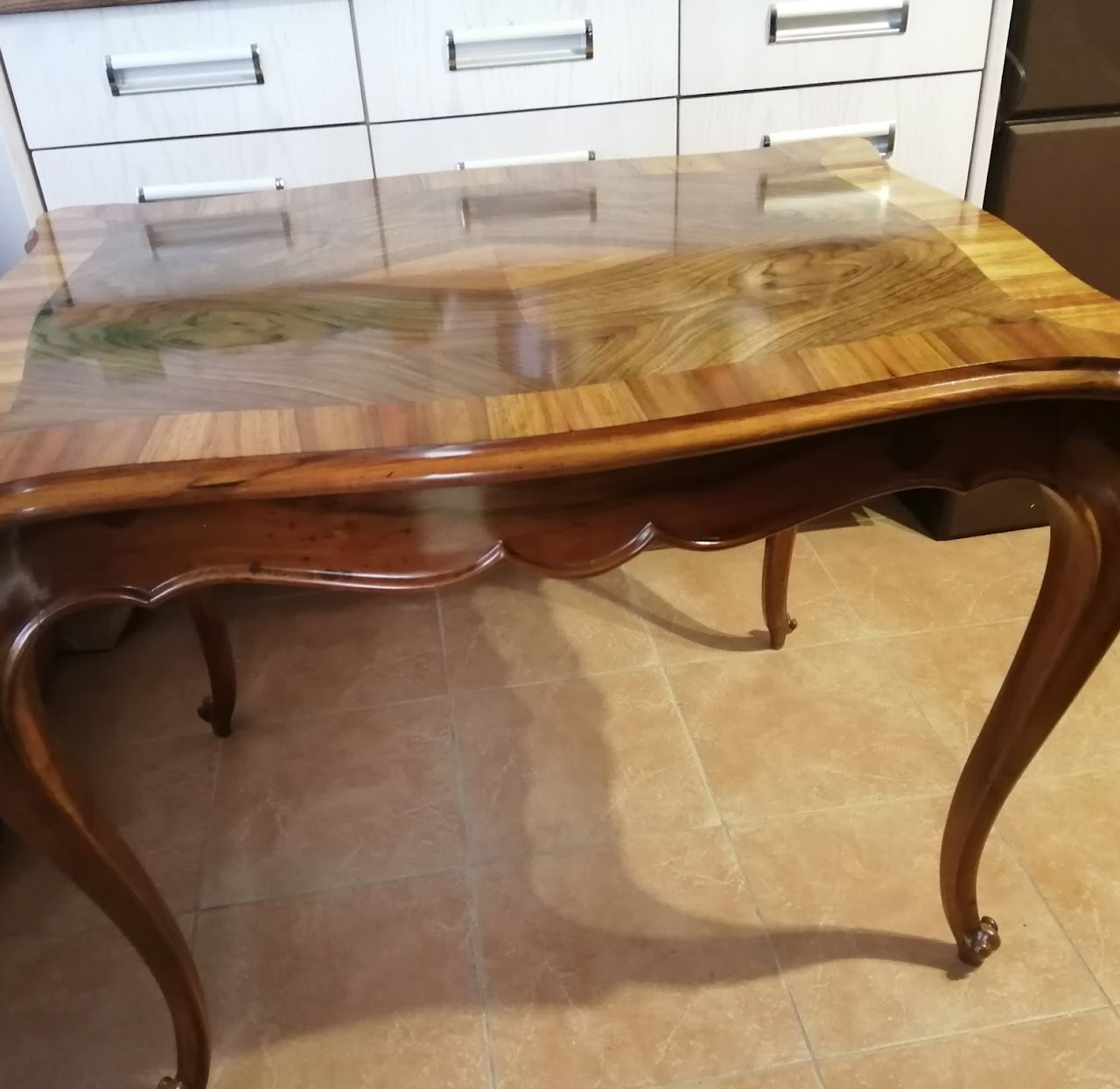 Wooden table with a polished surface and curved legs, situated on a tiled floor with cabinets in the background.