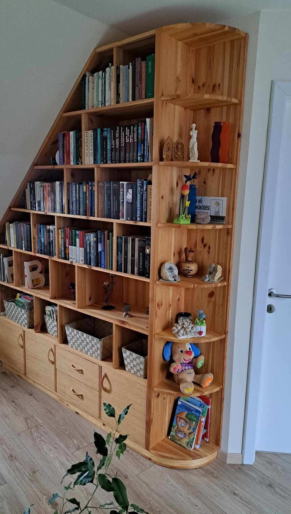 Corner wooden bookshelf filled with books, decorative items, and children's toys, with a closed white door to the right and a plant in the lower left corner.