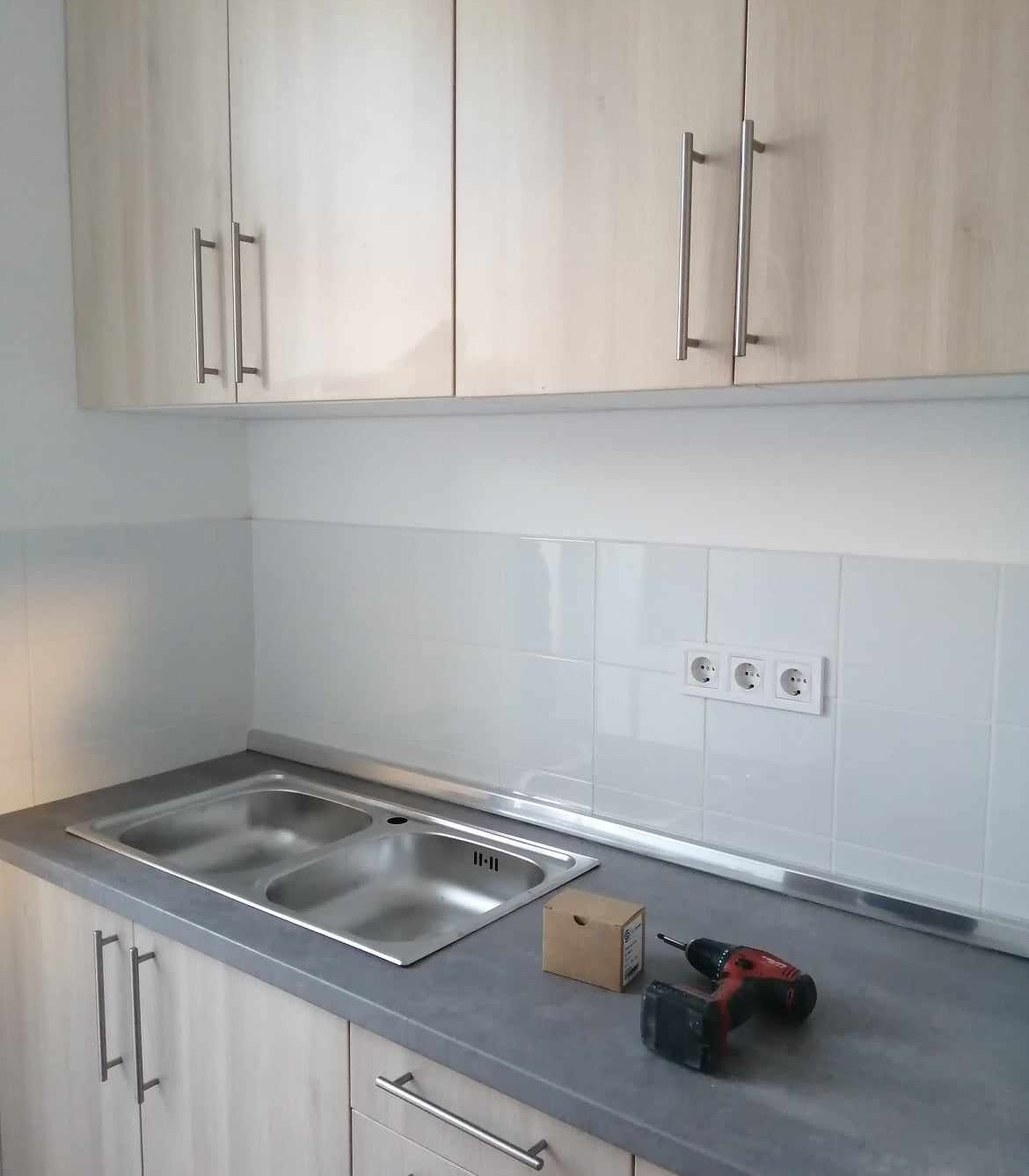 Kitchen countertop with a mounted double sink, a cordless drill, a small cardboard box, and an electrical outlet on the tiled wall.