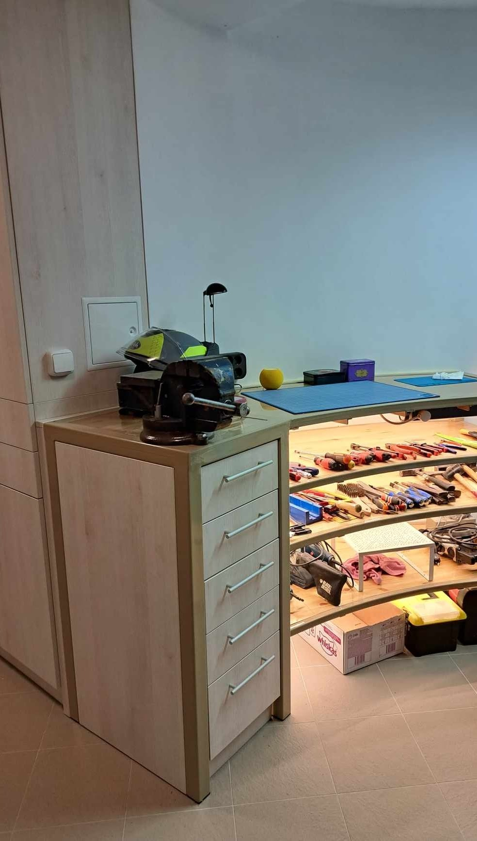 A workplace with woodworking tools, including screwdrivers, pliers, and other hand tools, displayed on a curved wooden shelf. There are various small items and equipment on a light-colored wooden cabinet, with a box labeled 'Whiskas' on the floor underneath. The background features a white wall and a workspace with a blue mat.