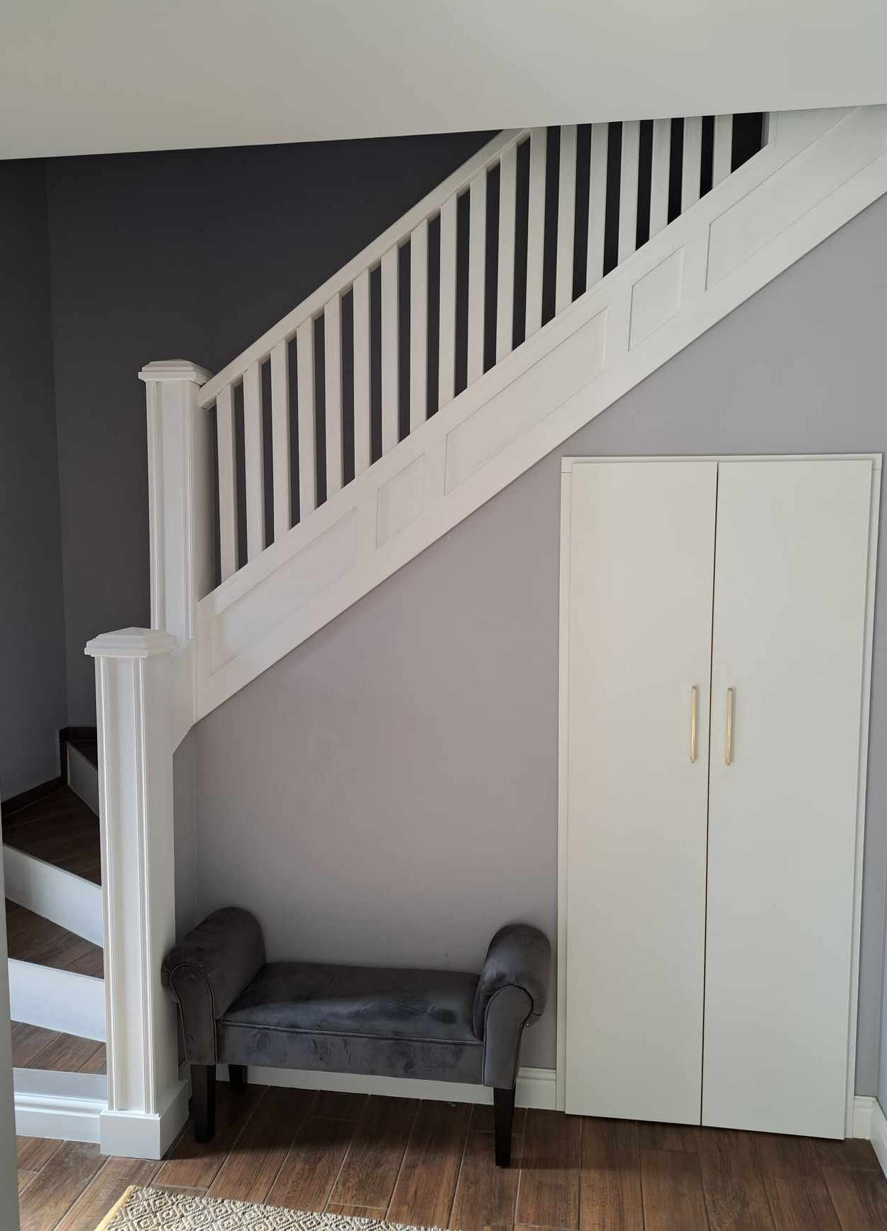 Inside view of a home with a staircase, black and white railings, a small gray velvet bench, a tall white cabinet with gold handles, and wood flooring.