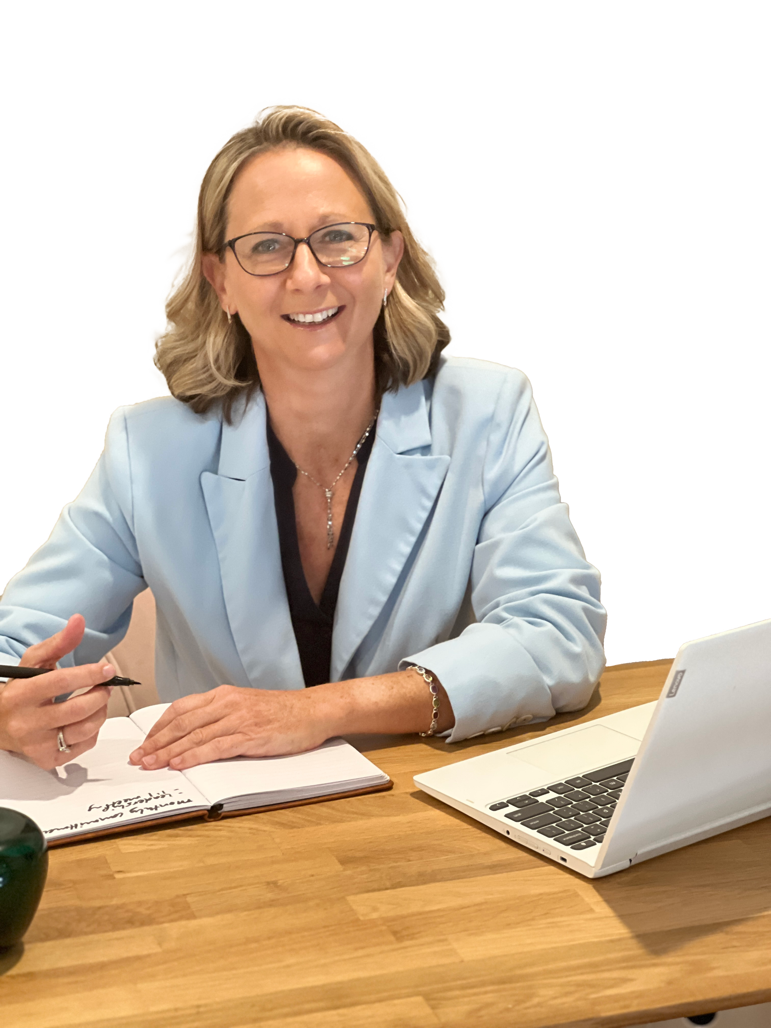 Smiling woman with blonde hair, glasses, wearing a light blue blazer, sitting at a wooden desk with a laptop, notebook, and pen.