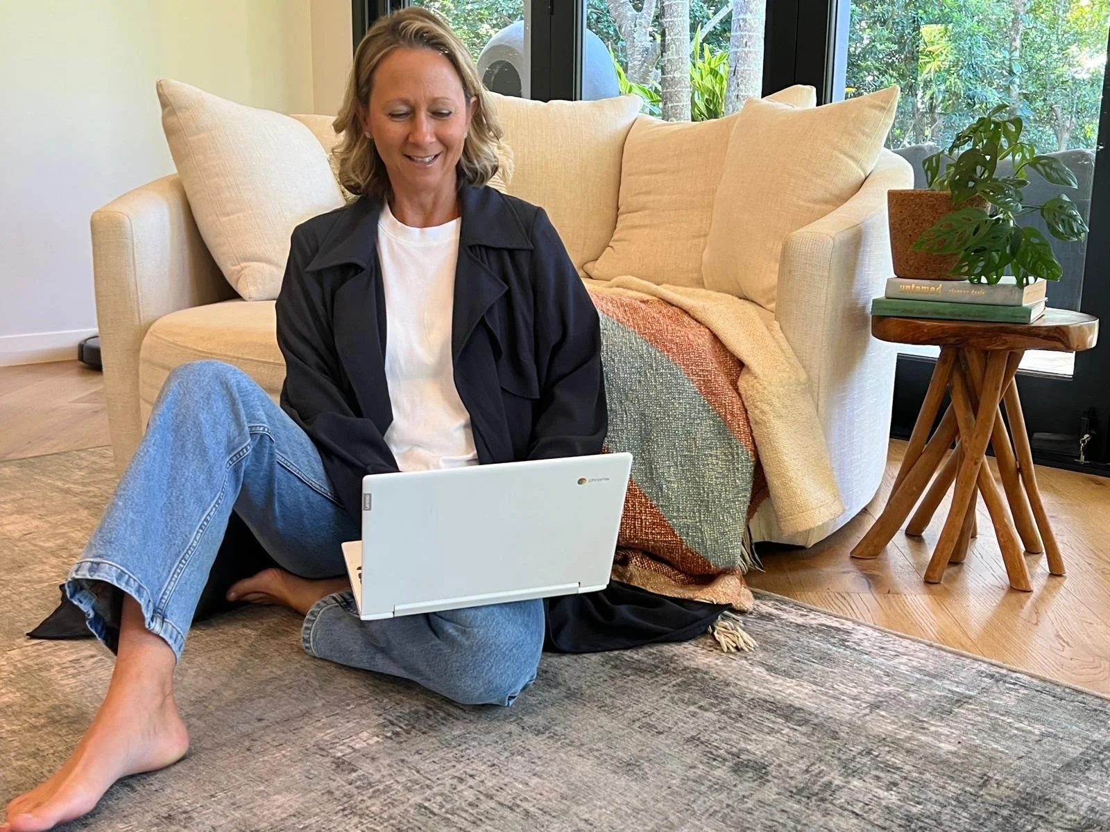 A woman with blonde hair, wearing a white t-shirt and black jacket, sitting on the floor with legs crossed, looking at a laptop, in a cozy living room with a beige sofa, pillows, a wooden side table with books and a potted plant, large window with greenery outside, and a rug.