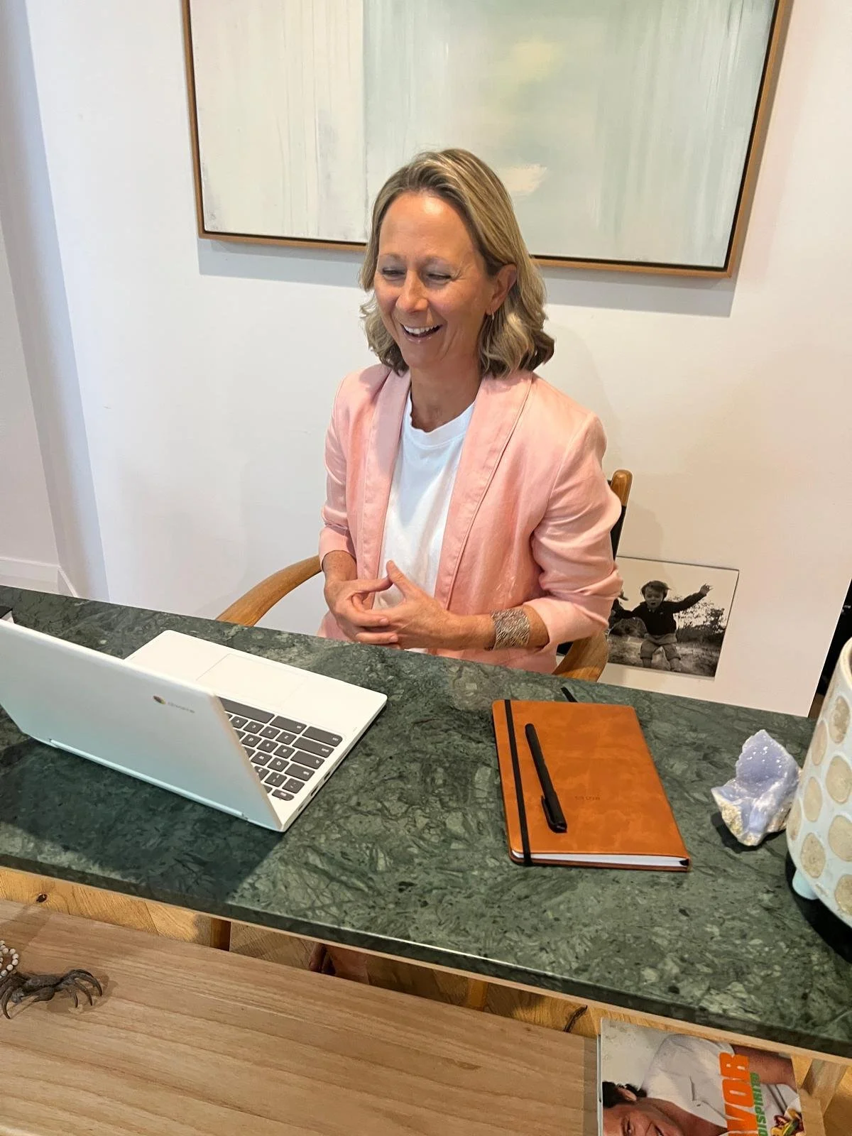 A woman with shoulder-length blonde hair wearing a pink blazer and white shirt, smiling and sitting at a green marble table with a laptop, a brown notebook with a black pen, and a decorative item on the table.
