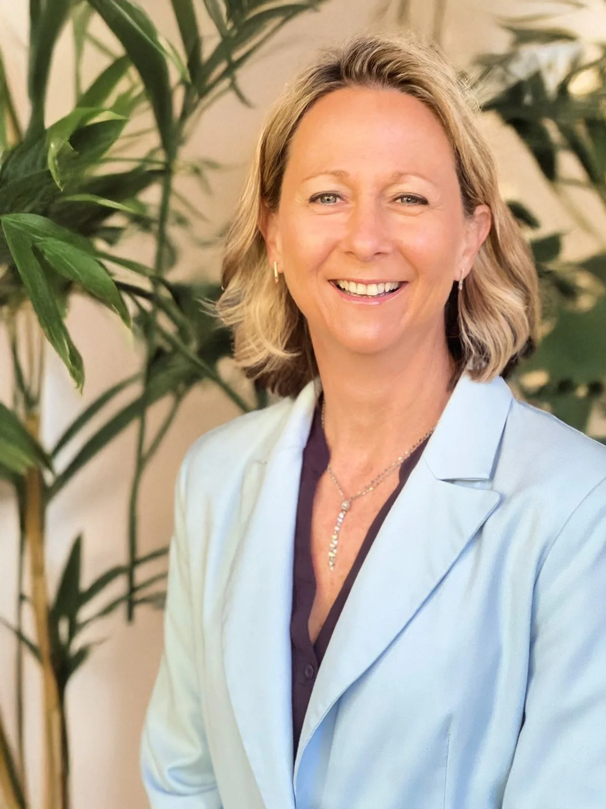 A smiling woman with shoulder-length blonde hair, wearing a light blue blazer and dark blouse, standing in front of green leafy plants.