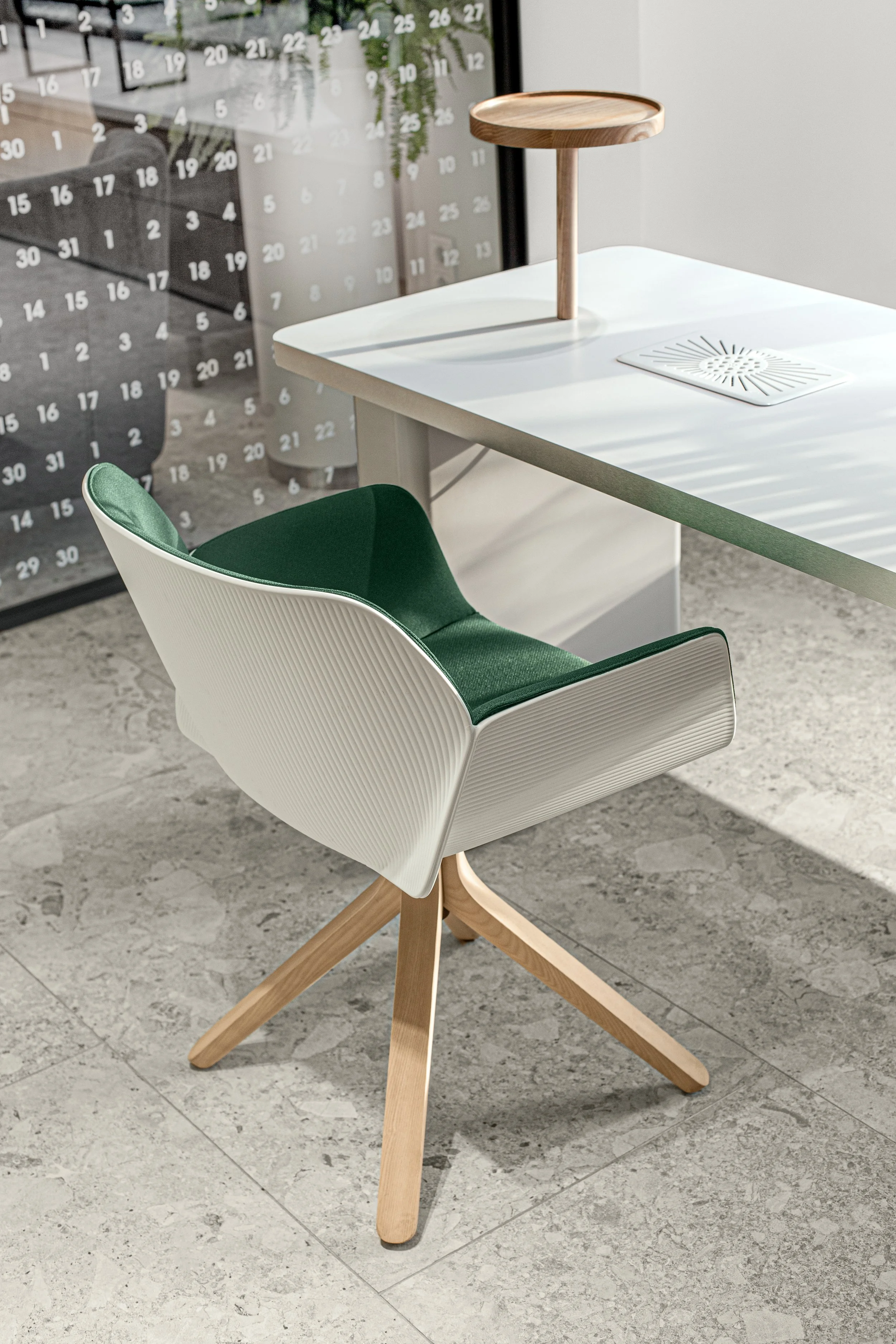 Modern office chair with a white exterior shell and green cushion, located next to a white desk with a small wooden table on top, in a bright room with a gray textured floor and a glass wall showing a calendar and other office furniture.