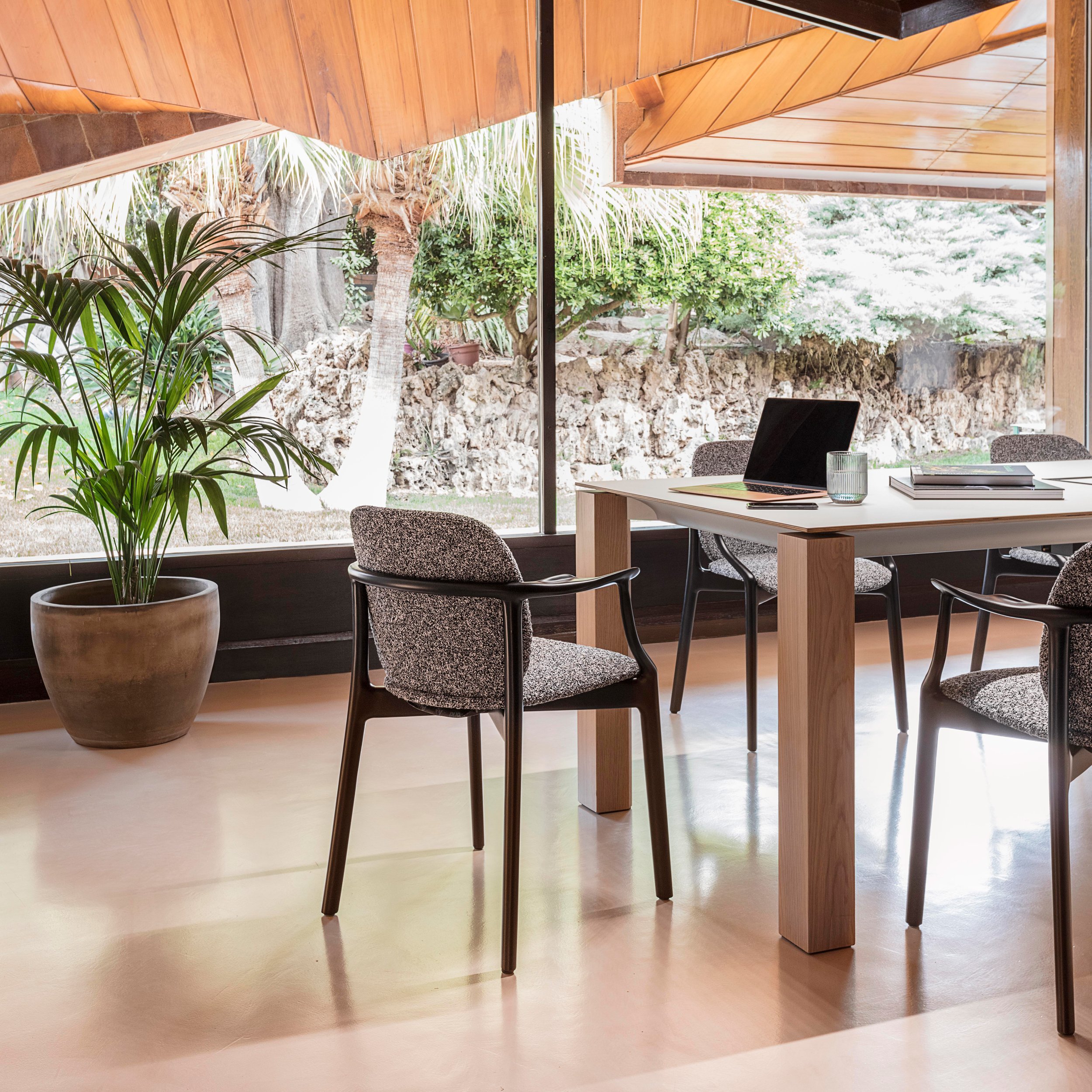 Indoor cafe with a small black table set for a meal, four pink chairs, two glasses of juice, and a potted plant, positioned near large floor-to-ceiling windows with a street view outside, and wooden shelves filled with various items on the left side.