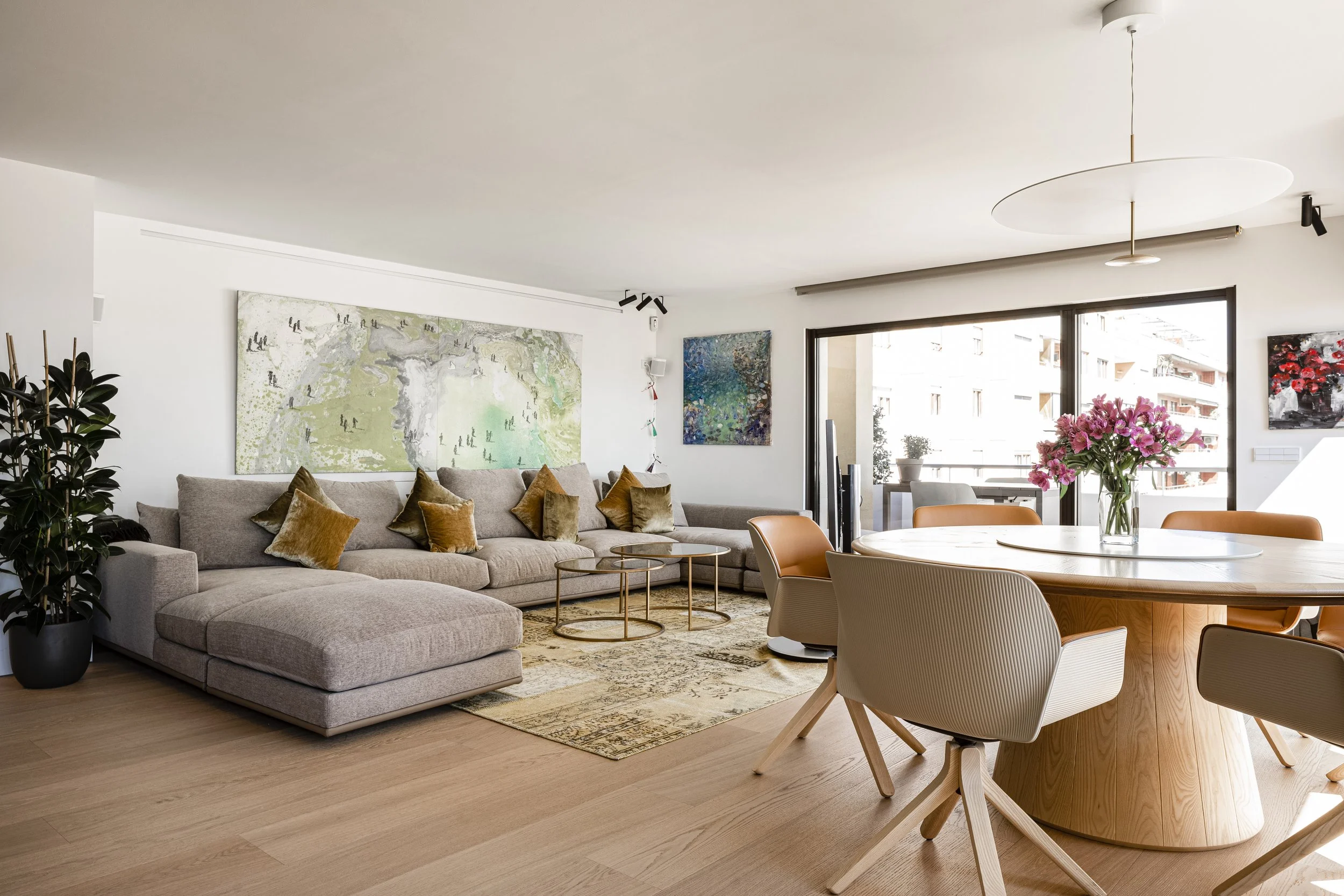A modern living room and dining area with a large sectional sofa, colorful pillows, abstract artwork, a round wooden dining table with beige and orange chairs, a tall glass vase with pink flowers, and a large sliding glass door leading to a balcony with city views.