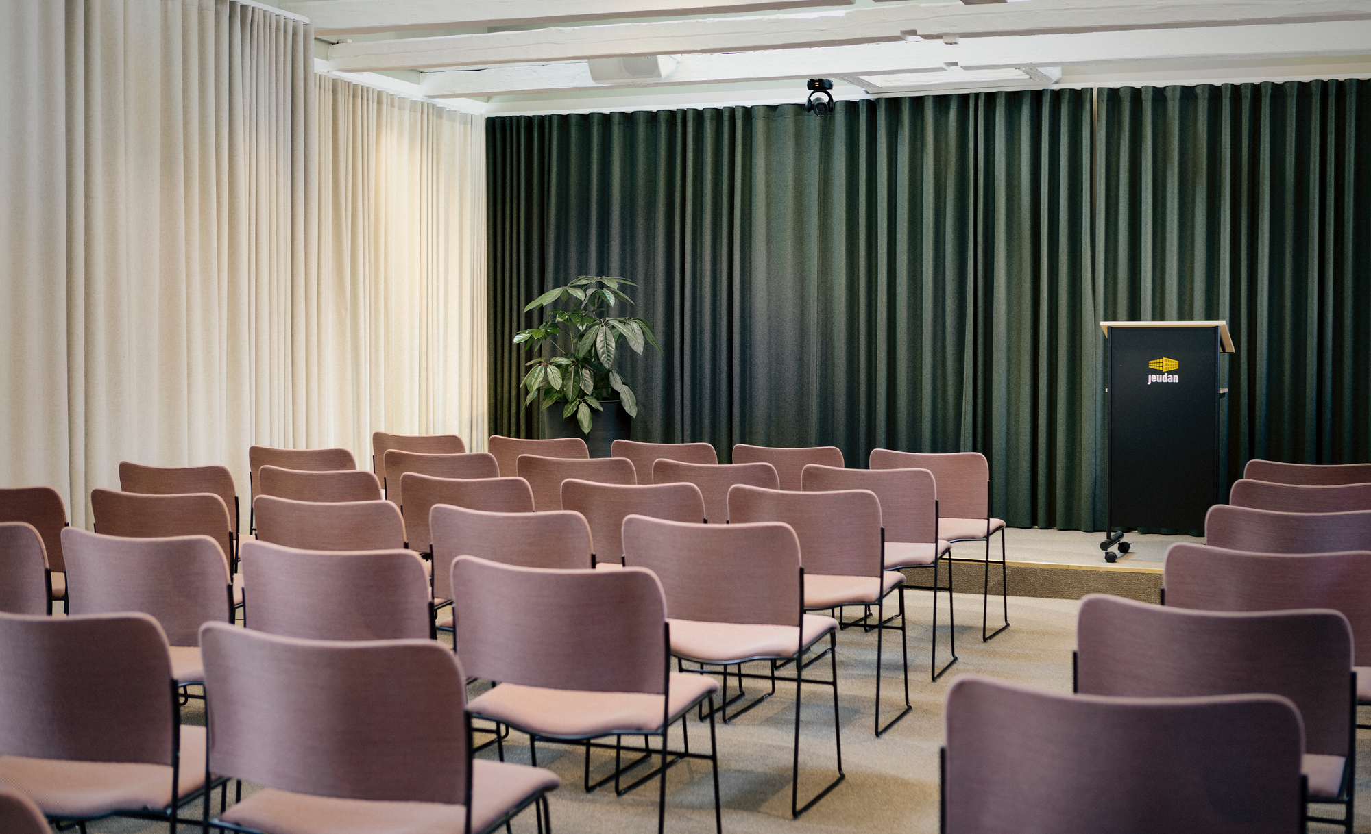 A modern conference room with a large wooden table, eight beige upholstered rolling chairs, three geometric pendant lights, large windows with a city view, and a whiteboard on a stand.