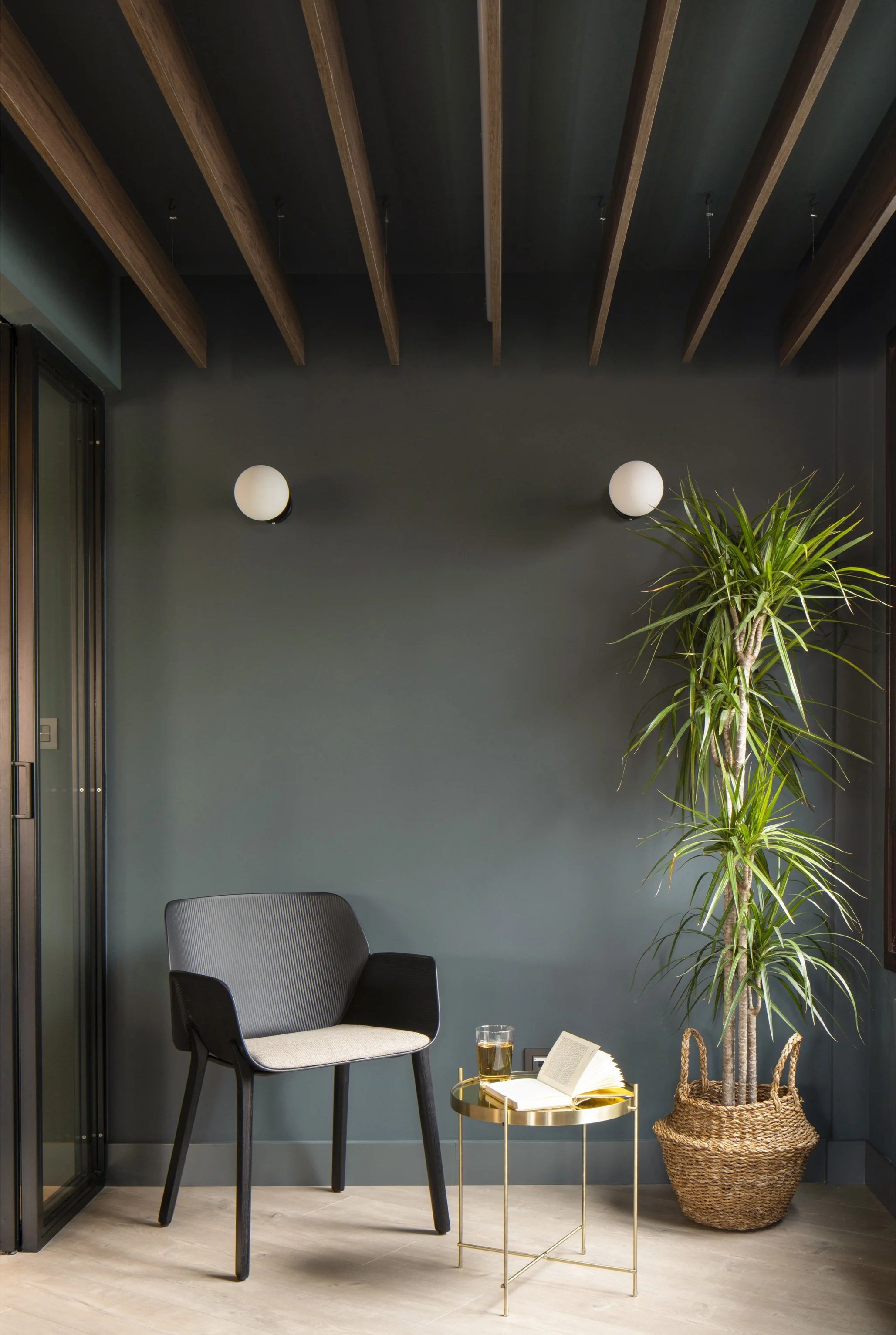 A modern interior corner with a black armchair, a small gold side table holding an open book and a glass of drink, a tall green potted plant in a woven basket, and two round wall lights on a dark green wall ceiling with exposed wooden beams.