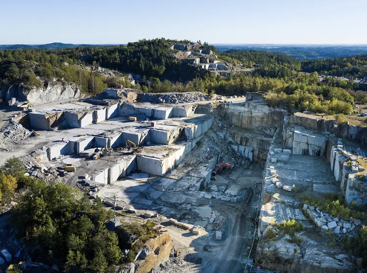 An aerial view of a large open-pit marble quarry with terraced extraction levels, bulldozers, and trucks, surrounded by green trees and distant hills.