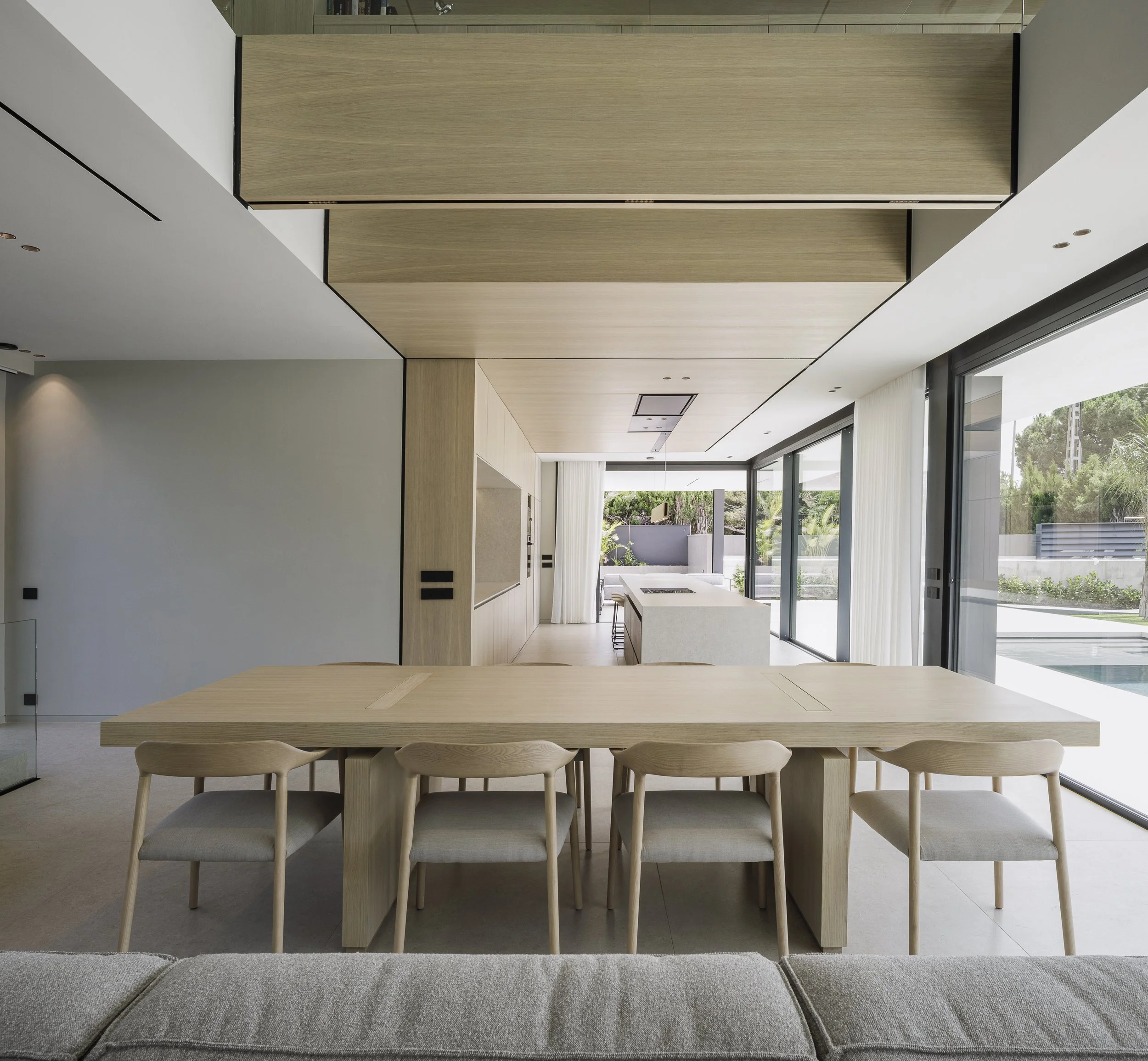 Modern restaurant with large windows, white ceiling, beige chairs around round tables set with white bowls and glasses, and large potted plants for decoration.