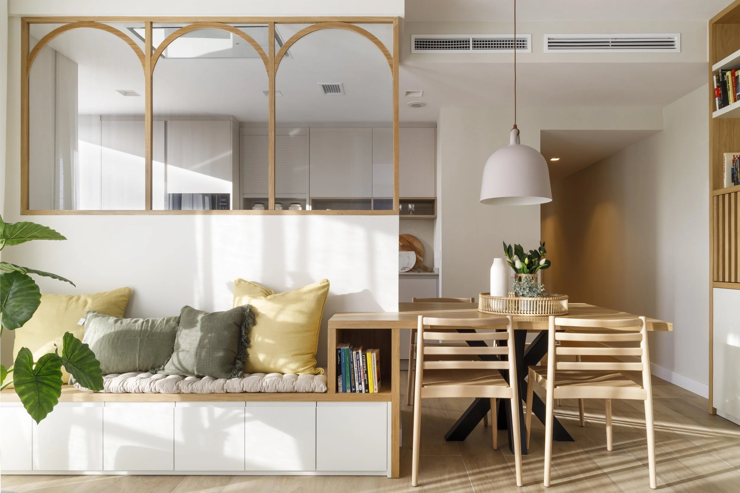 Living and dining area in a modern, minimalistic home with a white and wood color scheme, including a sofa with cushions, a round dining table with wooden chairs, and decorative vases and books.