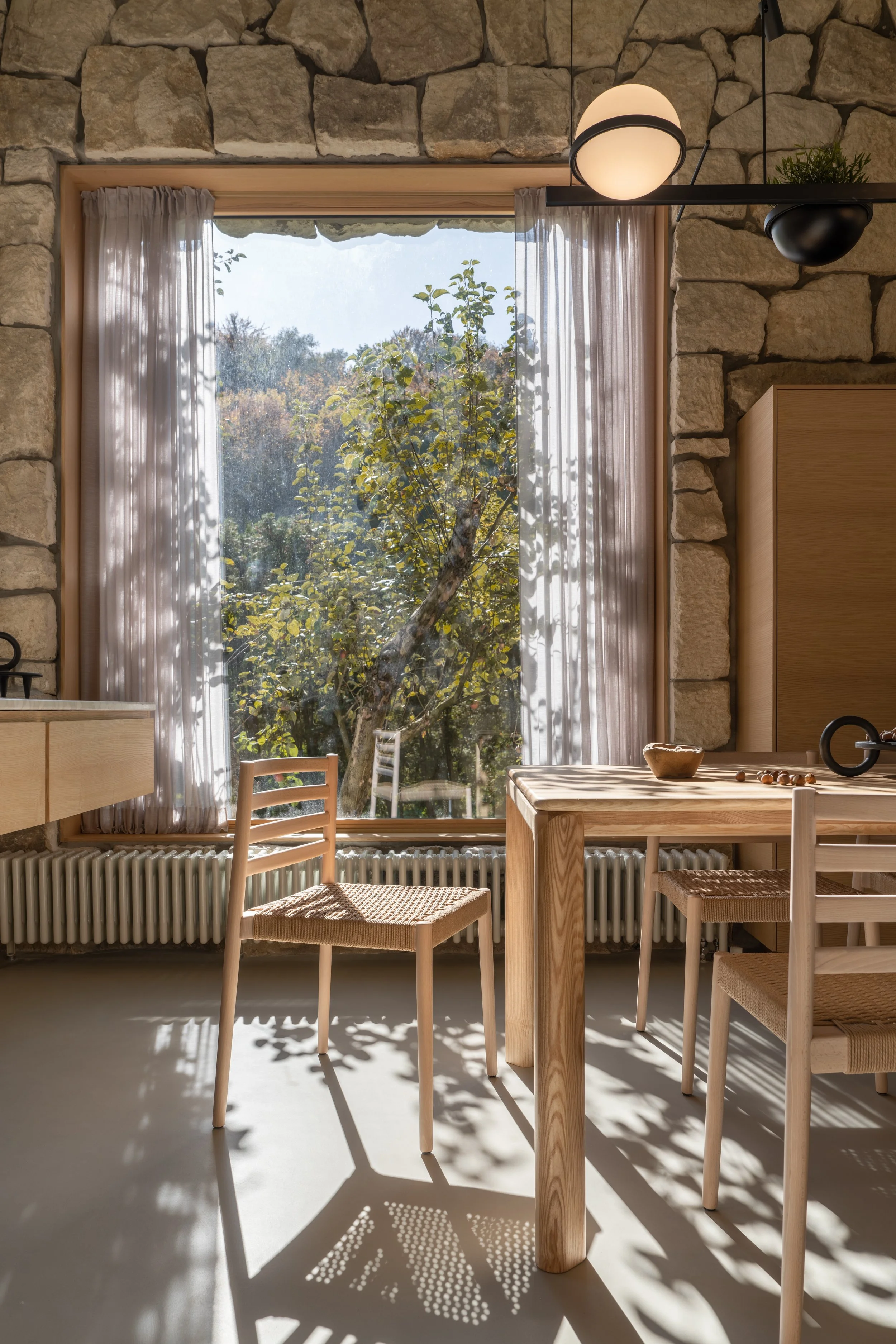 A cozy dining area with a wooden table and matching chairs in front of a large window with sheer curtains. Outside, a tree is visible in a sunny outdoor scene, and sunlight creates shadows on the floor inside.