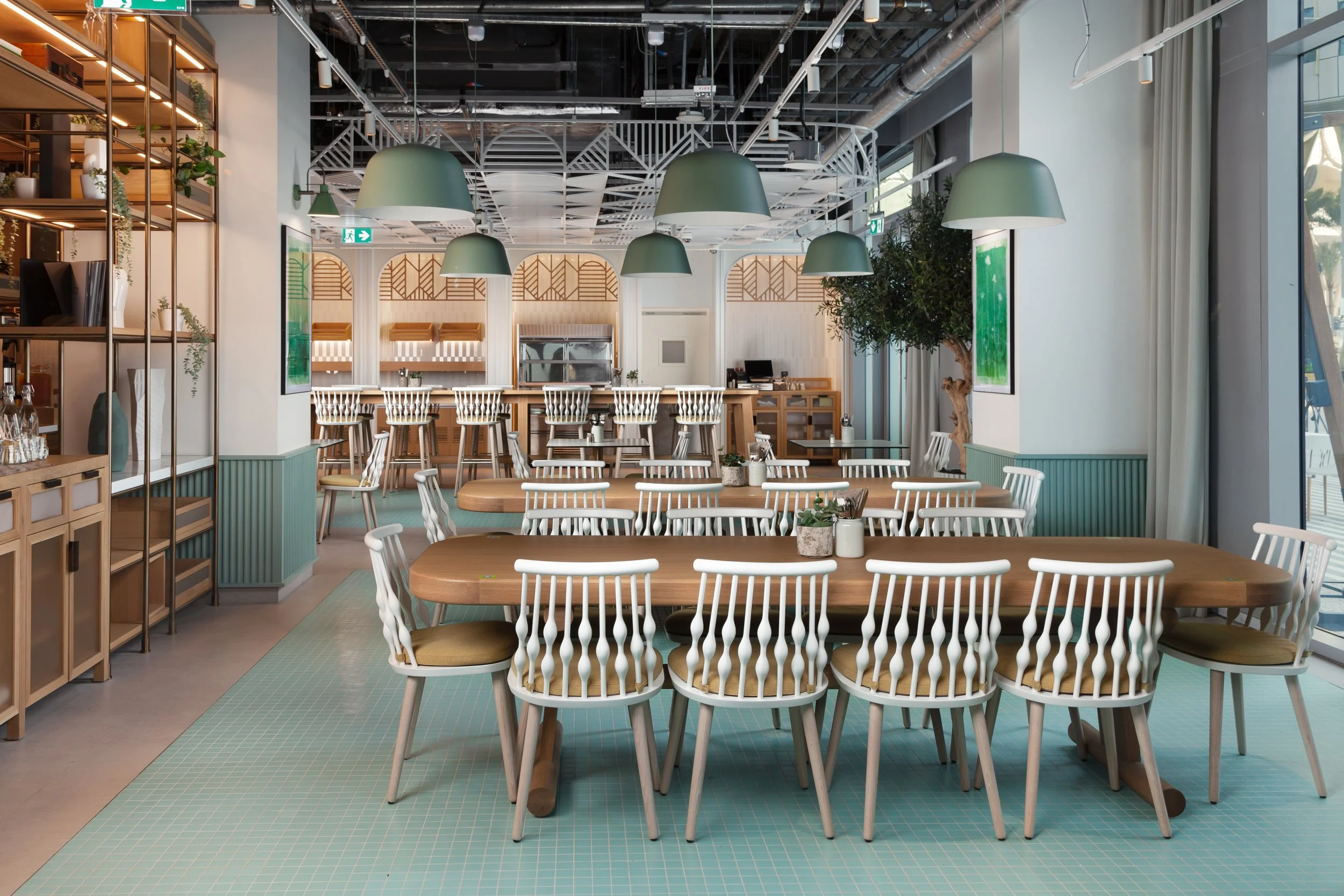 A cozy modern restaurant or cafe interior with a large wooden dining table surrounded by white chairs with beige cushions, green pendant lamps hanging from the ceiling, and a bar area at the back. There are decorative plants and large windows letting in natural light.
