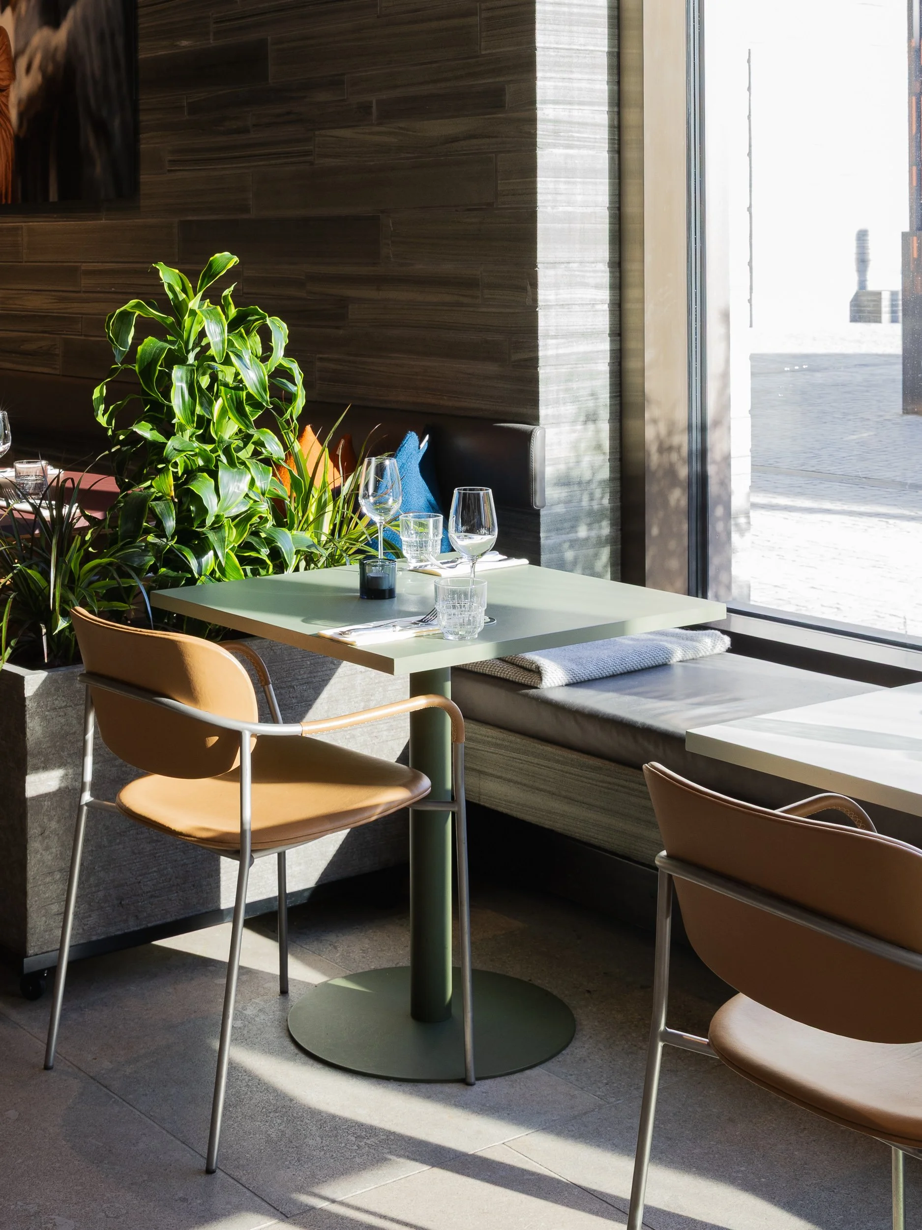 A cozy restaurant corner with a table set for two, featuring wine glasses, water glasses, and candles, next to large window with natural sunlight.