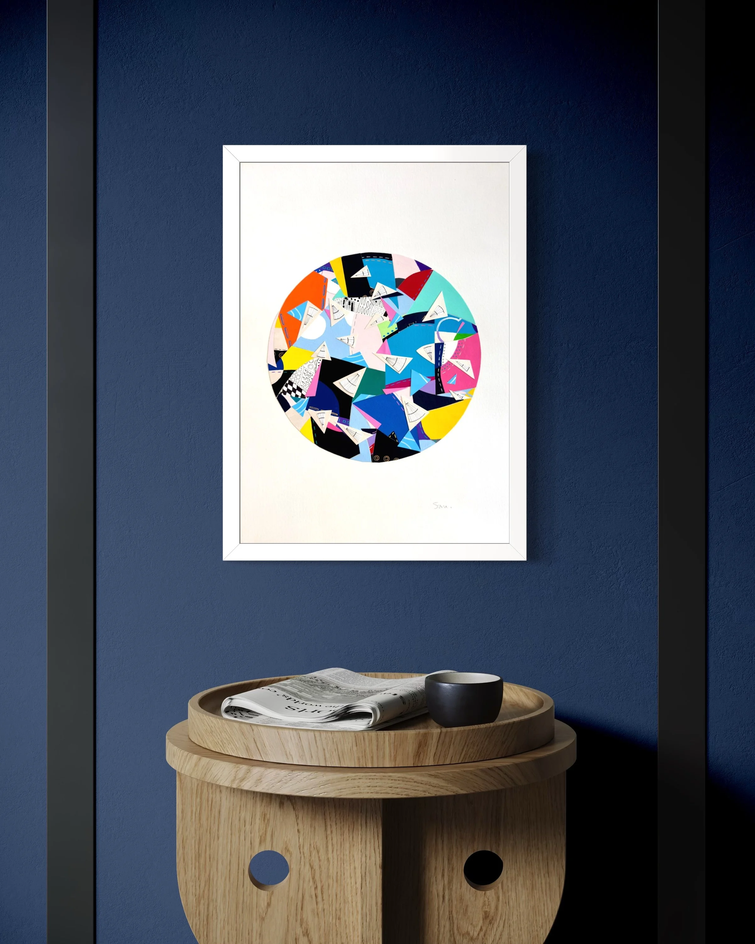 A Restless Centre, A3 abstract mixed media collage with a circular centre of coloured fragments and triangular pen drawings displayed framed on a blue wall in a modern living room.