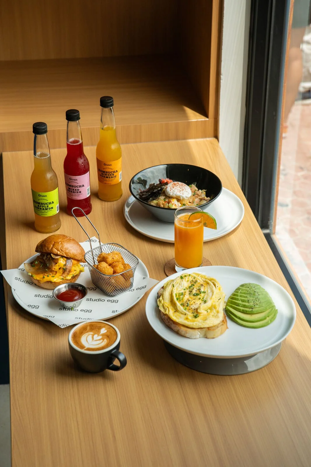 Studio Egg Ubud Bali menu spread — Velvet Egg Avocado, Egg Sandwich with tater tots, Oyakodon with poached egg on Japanese rice, kombucha bottles and cappuccino