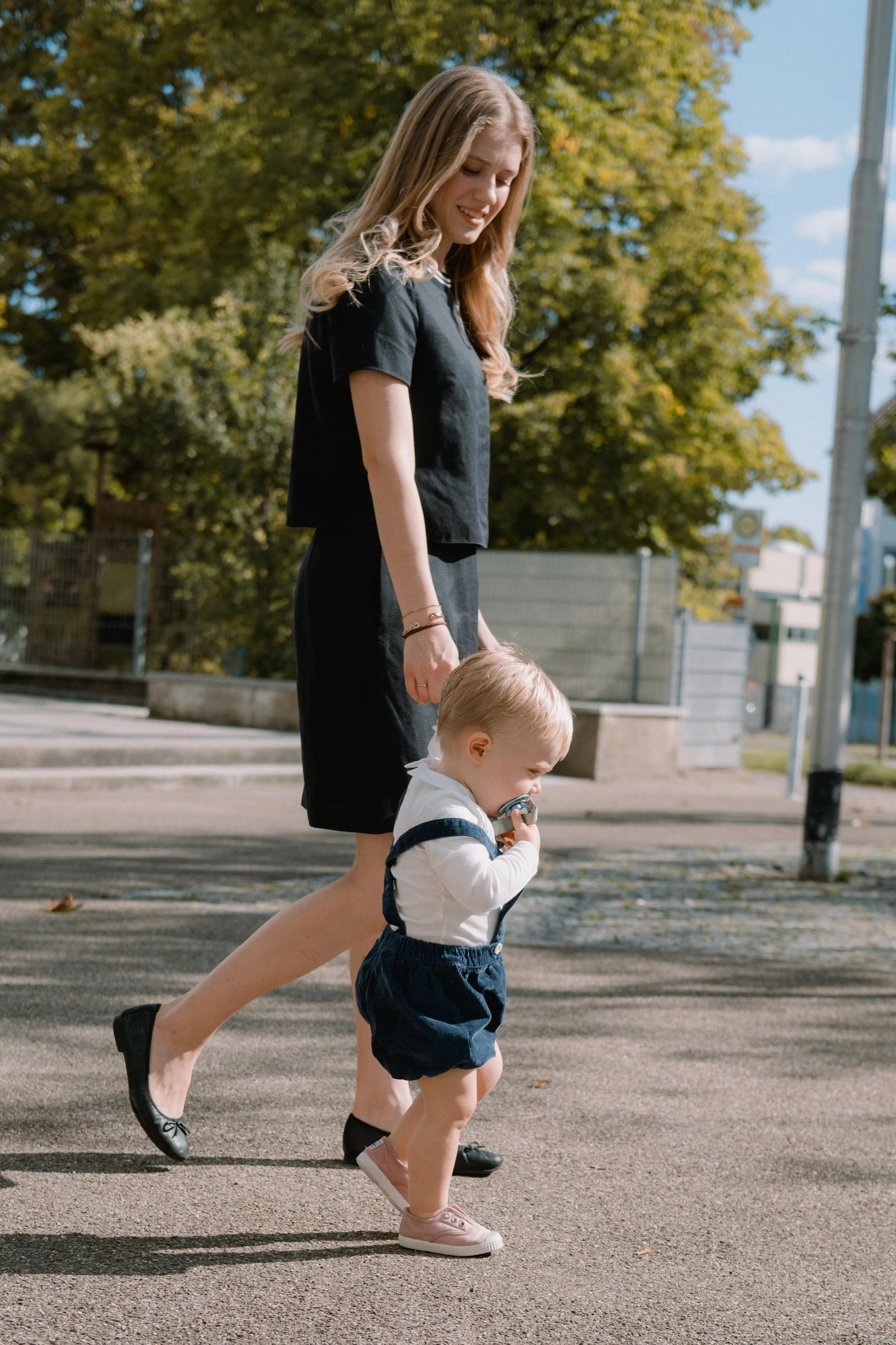 Eine junge Frau und ein kleines Kind, die auf einer Straße im Freien spazieren gehen. Die Frau trägt ein schwarzes Kleid und Schuhe, das Kind trägt einen weißen Pullover, dunkle Shorts und rosa Schuhe. Im Hintergrund sind Bäume mit herbstlichem Laub 