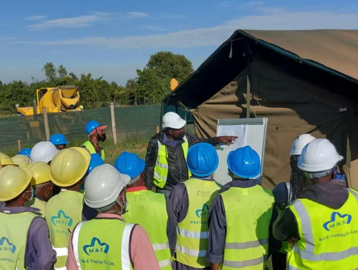 Group of construction workers wearing safety helmets and vests, listening to a supervisor giving instructions outdoors near a fence, with construction equipment and trees in the background.
