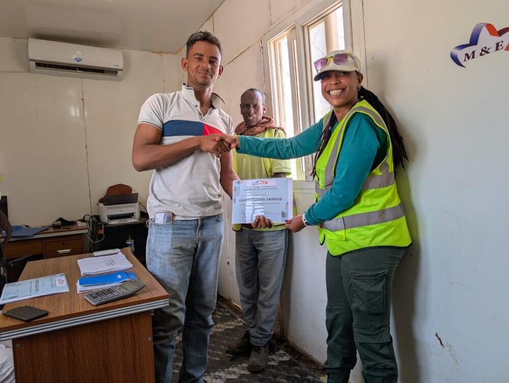 A woman in a yellow safety vest hands a certificate to a young man, shaking hands inside an office with two other people watching. The woman is smiling broadly, and the office has papers, a calculator on a desk, and a window in the background.