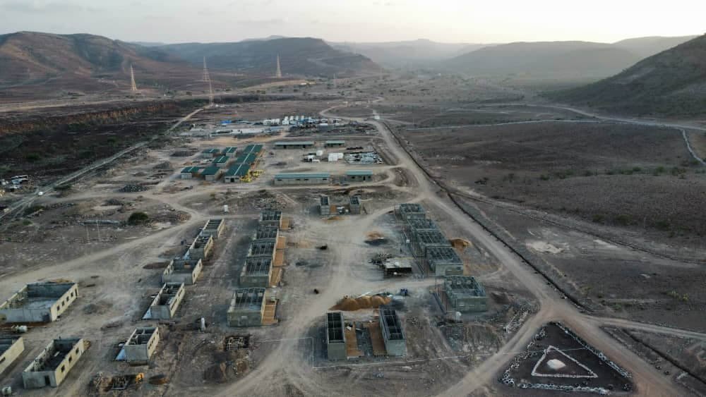 Aerial view of a construction site in a desert area with mountains in the background, showing multiple partially built structures and construction materials.
