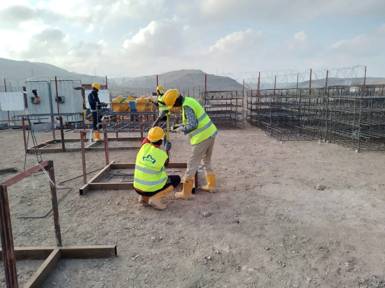 Construction workers wearing yellow safety vests and helmets working on metal frameworks outdoors with mountains in the background.