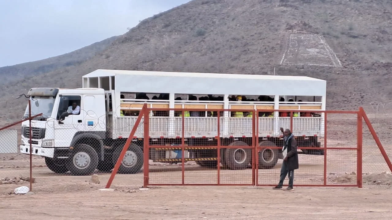 A large white truck with open sides parked behind a red metal gate, with a man standing outside near the gate in a desert landscape, mountain in the background.