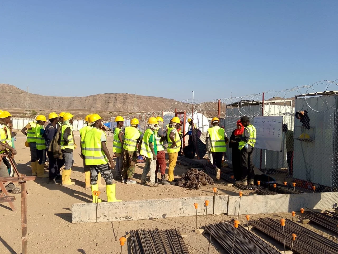 Construction workers wearing yellow safety helmets and reflective vests gathered on a construction site in a desert landscape. They are listening to a person giving instructions near a fenced area with barbed wire, construction materials, and a whiteboard.