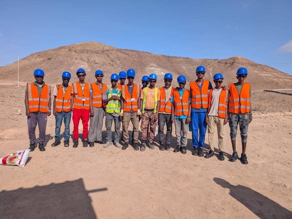A group of 13 construction workers wearing orange safety vests and blue helmets standing in a desert landscape with hills in the background. Some are wearing sunglasses and carrying notebooks.