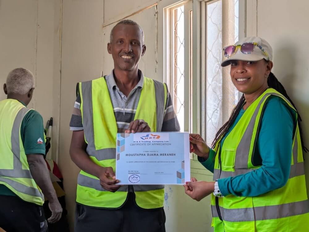 A man and a woman, both wearing reflective safety vests, are smiling and holding a certificate of appreciation. The man is wearing a striped polo shirt, and the woman is wearing a cap and sunglasses. They are indoors near a window, with two other peo