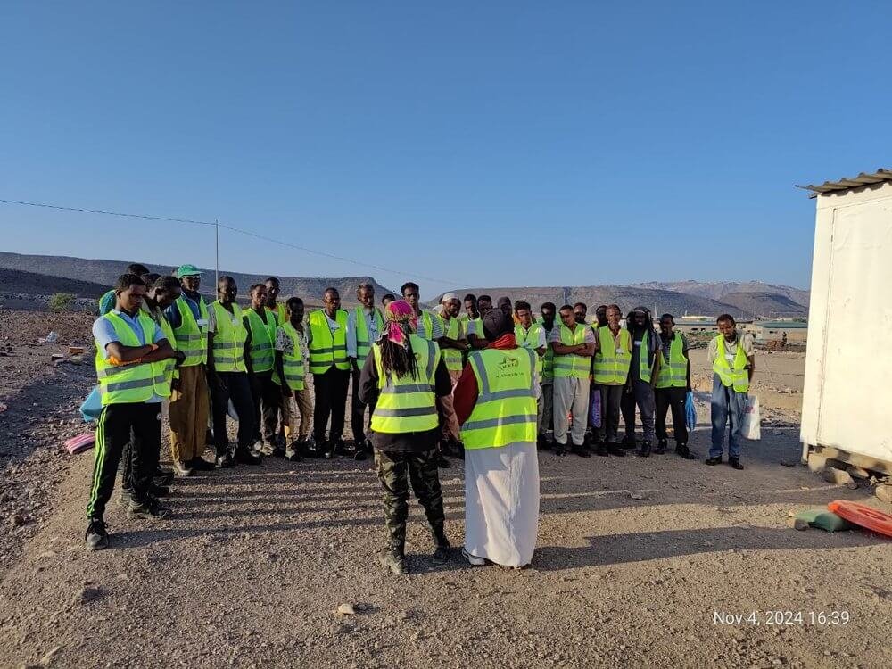 Group of people wearing yellow safety vests gathered outdoors on a dirt ground, with a clear blue sky and distant mountains in the background, during daytime.