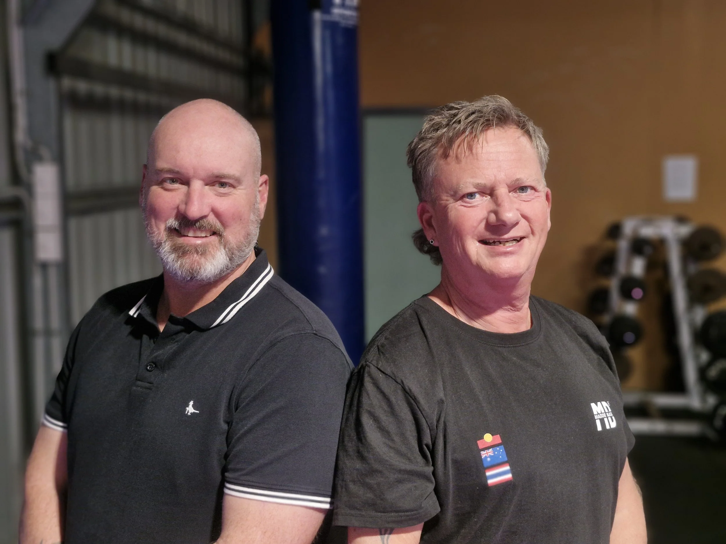 Two men standing side by side indoors, smiling at the camera. The man on the left has a bald head and a beard, wearing a black polo shirt. The man on the right has short, light hair and is wearing a black t-shirt with flags on it. Background includes gym equipment.