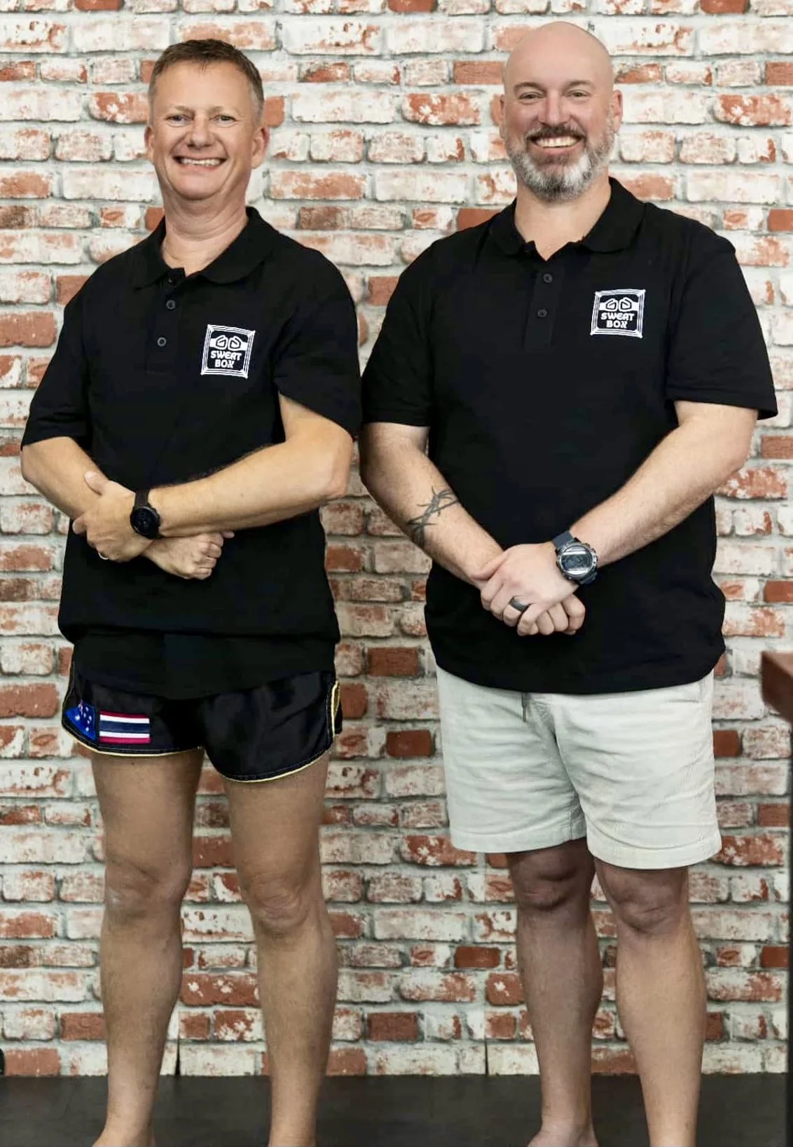 Two men stand side by side against a brick wall, smiling. Both are wearing black shirts with a logo that says 'Sweat Box.' The man on the left is wearing shorts with an American flag and a blue patch, while the man on the right is wearing beige shorts. They have their arms crossed or at their sides.