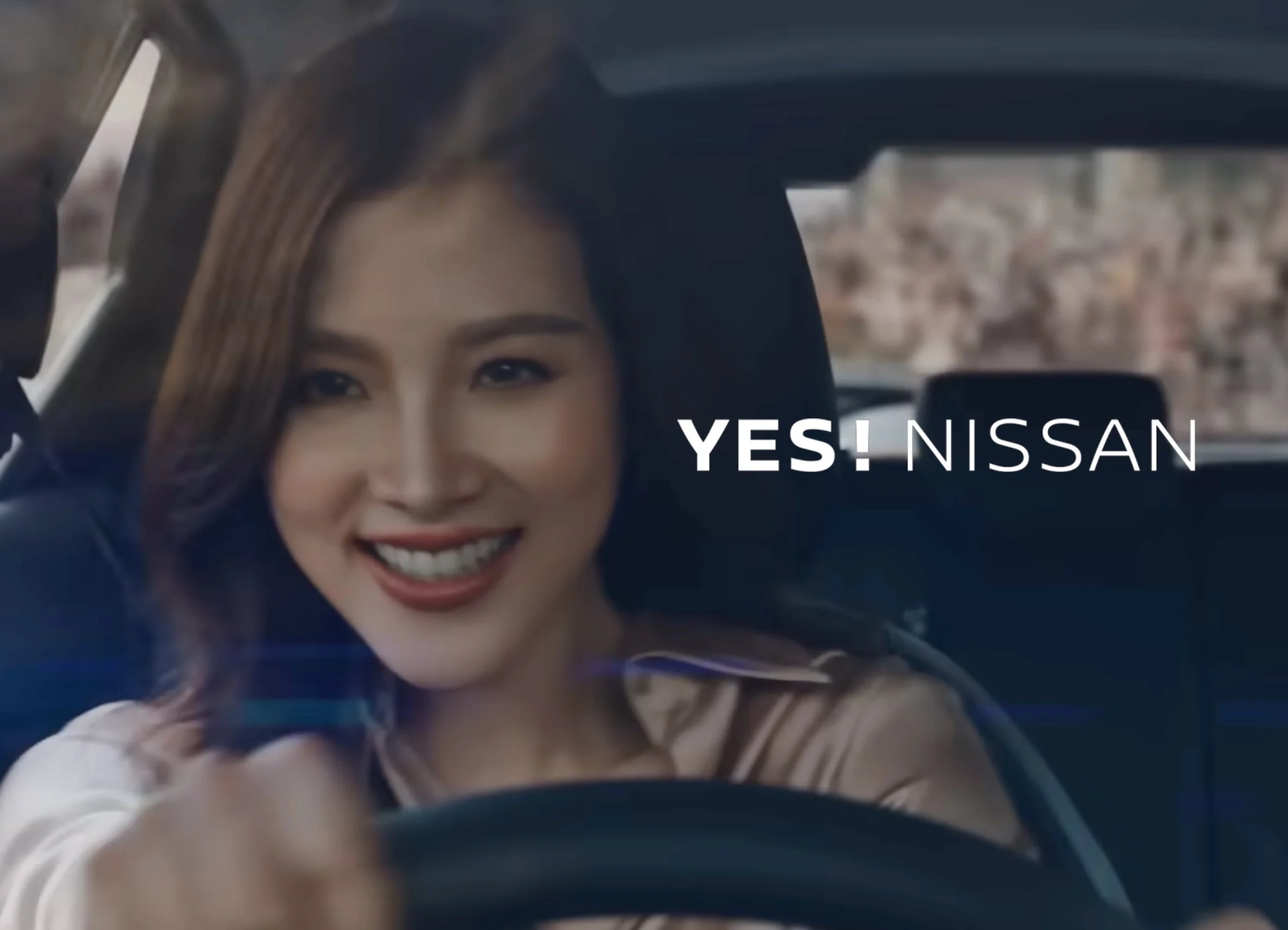 Close-up of a young woman with brown hair smiling from inside a vehicle, with part of her face and hand visible.