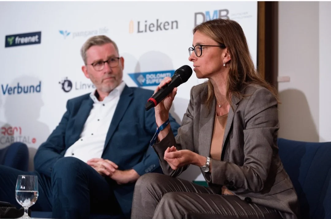 Iris Wagner bei einer Podiumsdiskussion, sie trägt eine Brille und einen Anzug und spricht ins Mikrofon, ein Mann in einem blauen Anzug hört zu, im Hintergrund Sponsorenlogos.