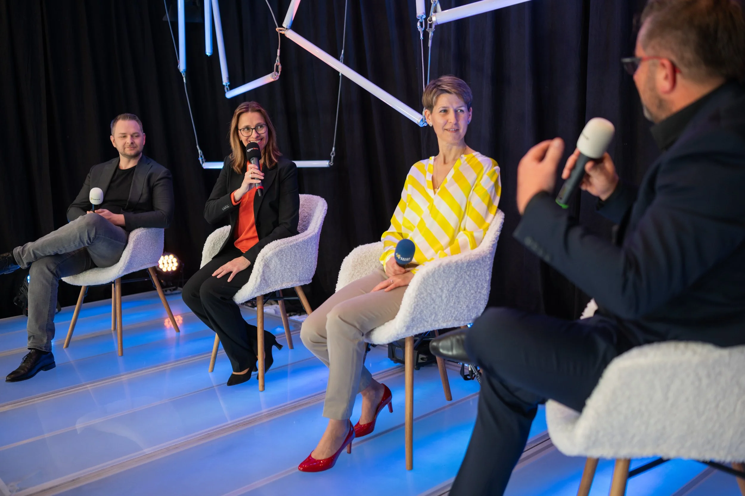 Vier Menschen, eine Person ist Iris Wagner, sitzen in einem Panel auf Stühlen, sprechen und halten Mikrofone, im Hintergrund dunkler Vorhang mit geometrischer neonbeleuchteter Dekoration.