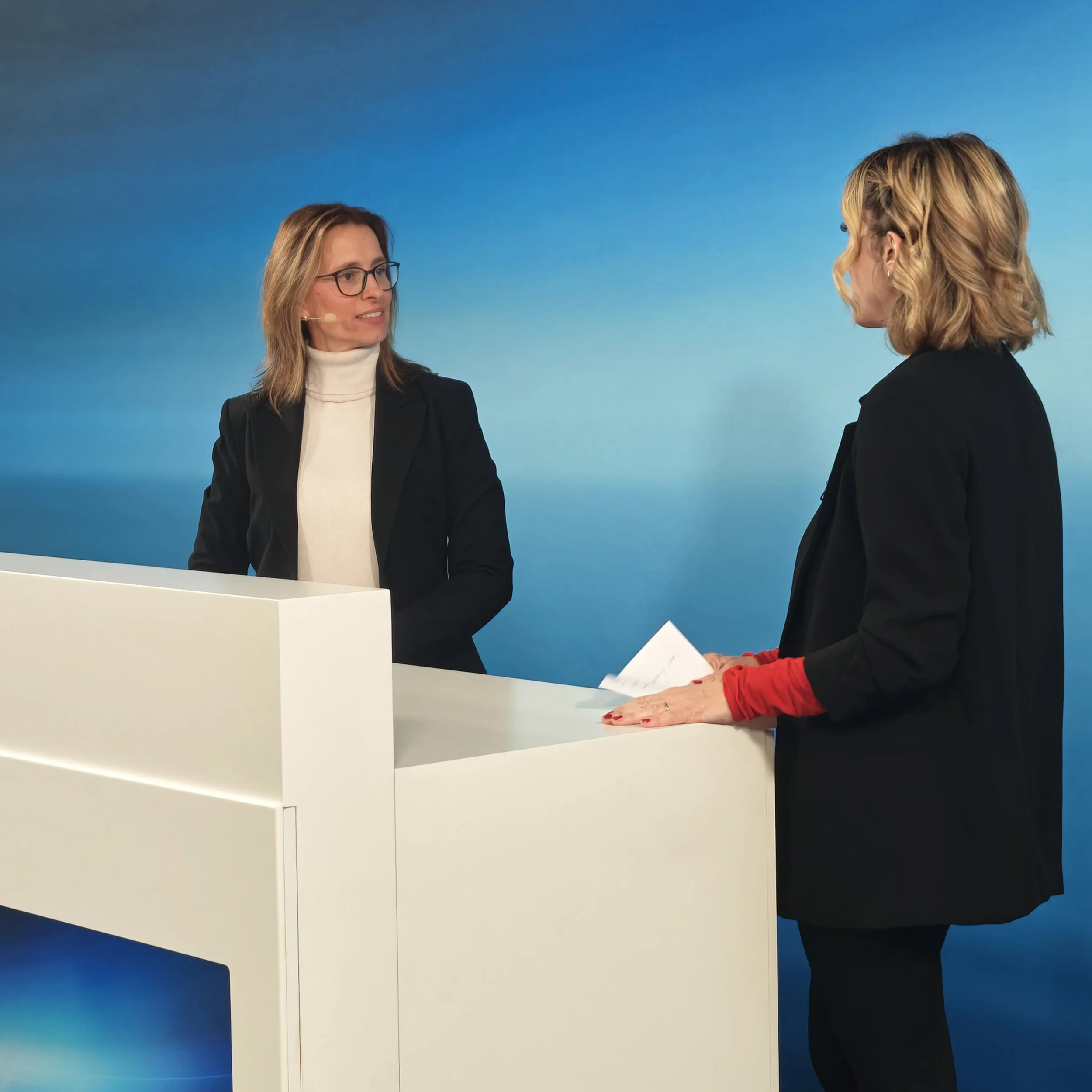 Iris Wagner im Gespräch mit der Moderatorin am Tresen, eine trägt eine schwarze Jacke mit roten Ärmeln, die andere eine schwarze Jacke, beide vor blauem Hintergrund.
