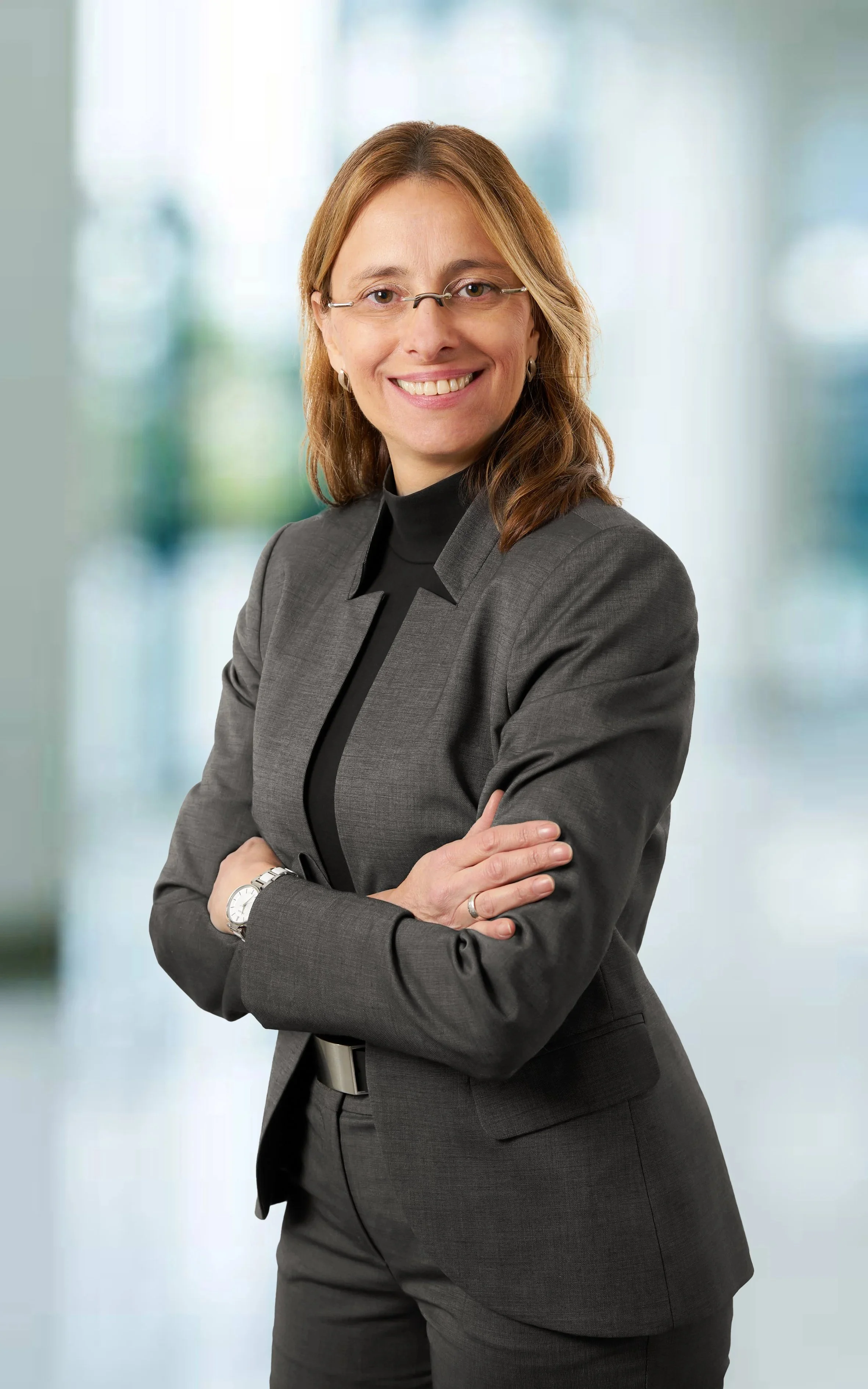 Iris Wagner in einem grauen Anzug mit Brille steht vor einem unscharfen Bürohintergrund.