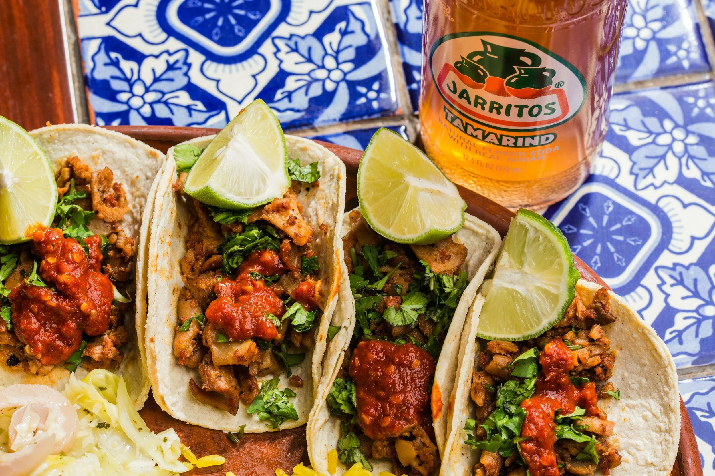 Three tacos filled with meat, topped with cilantro and salsa, garnished with lime wedges, on a wooden serving plate. A bottle of Jarritos tamarind soda in the background, on a blue patterned table.