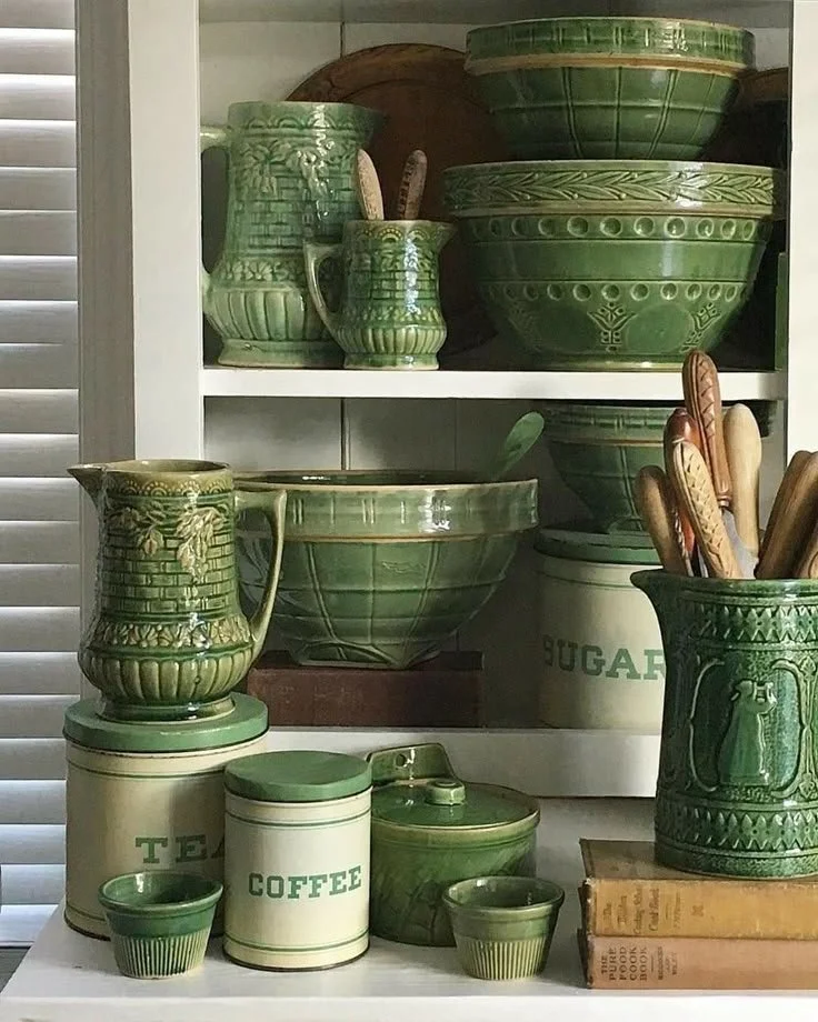 Green vintage ceramic kitchenware including bowls, pitchers, cups, a sugar container, and a coffee container, arranged on shelves and a table.