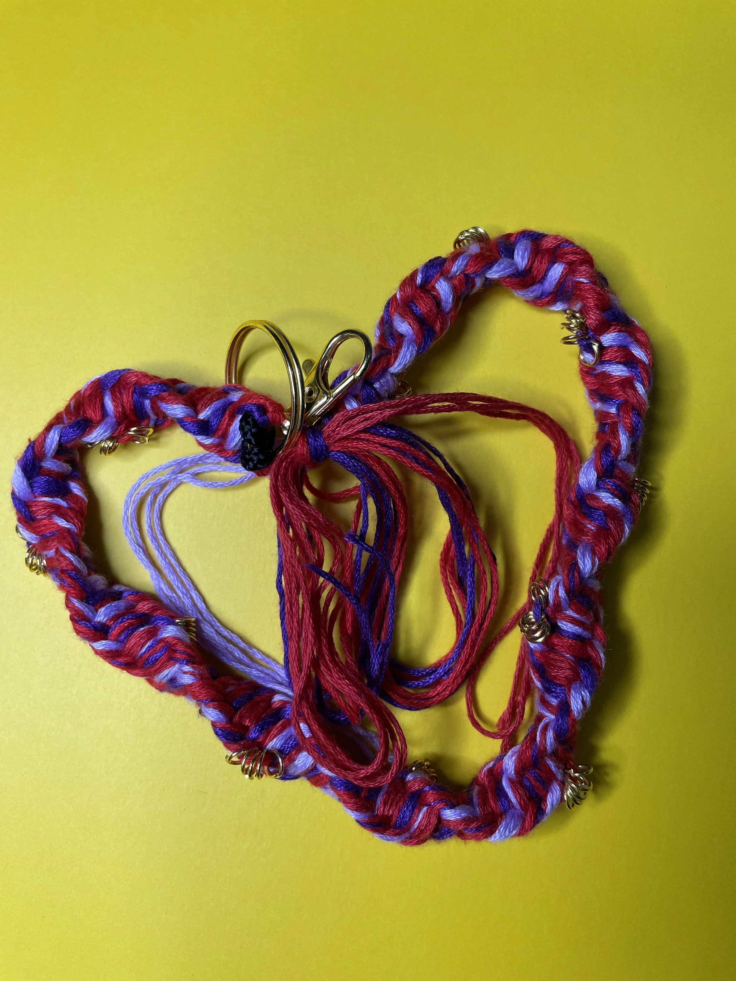 Colorful handmade friendship bracelet with red, purple, and lavender yarn, decorated with small metallic charms. The bracelet is lying on a bright yellow background.