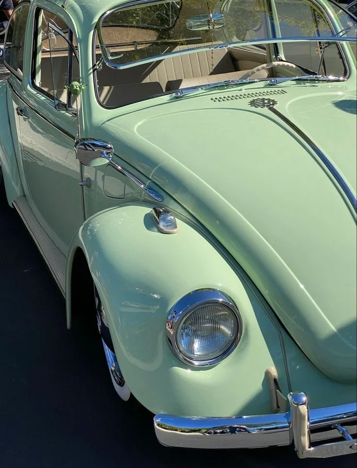 A vintage light green Volkswagen Beetle car parked outdoors, showing the front and side with chrome details and inside view of beige seats.