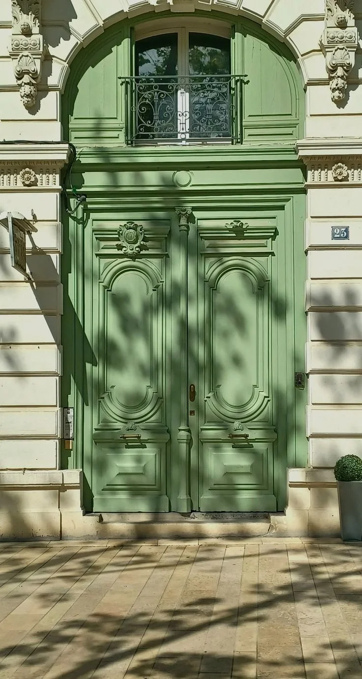 Green ornate double door with decorative molding, flanked by a cream-colored building and a small potted plant to the right, with shadows from nearby trees cast on the sidewalk.