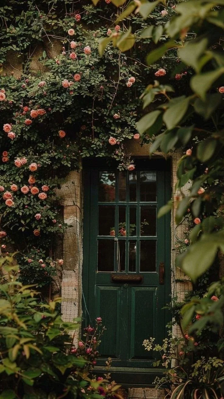 A green wooden door surrounded by lush flowering bushes with pink roses and green leaves, some of which partially obscure the view.