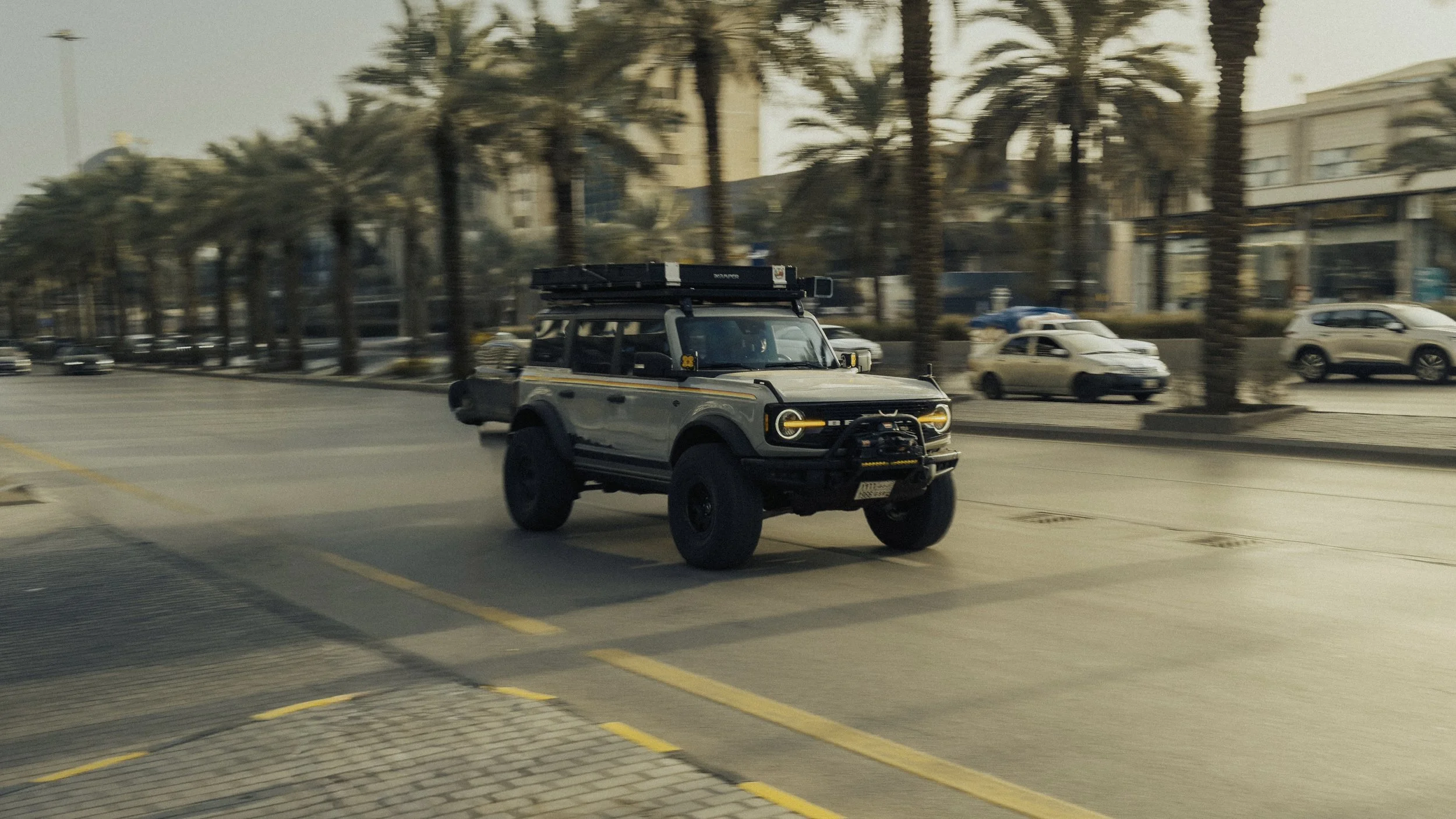 A black and white off-road vehicle driving on a city street lined with palm trees and cars.