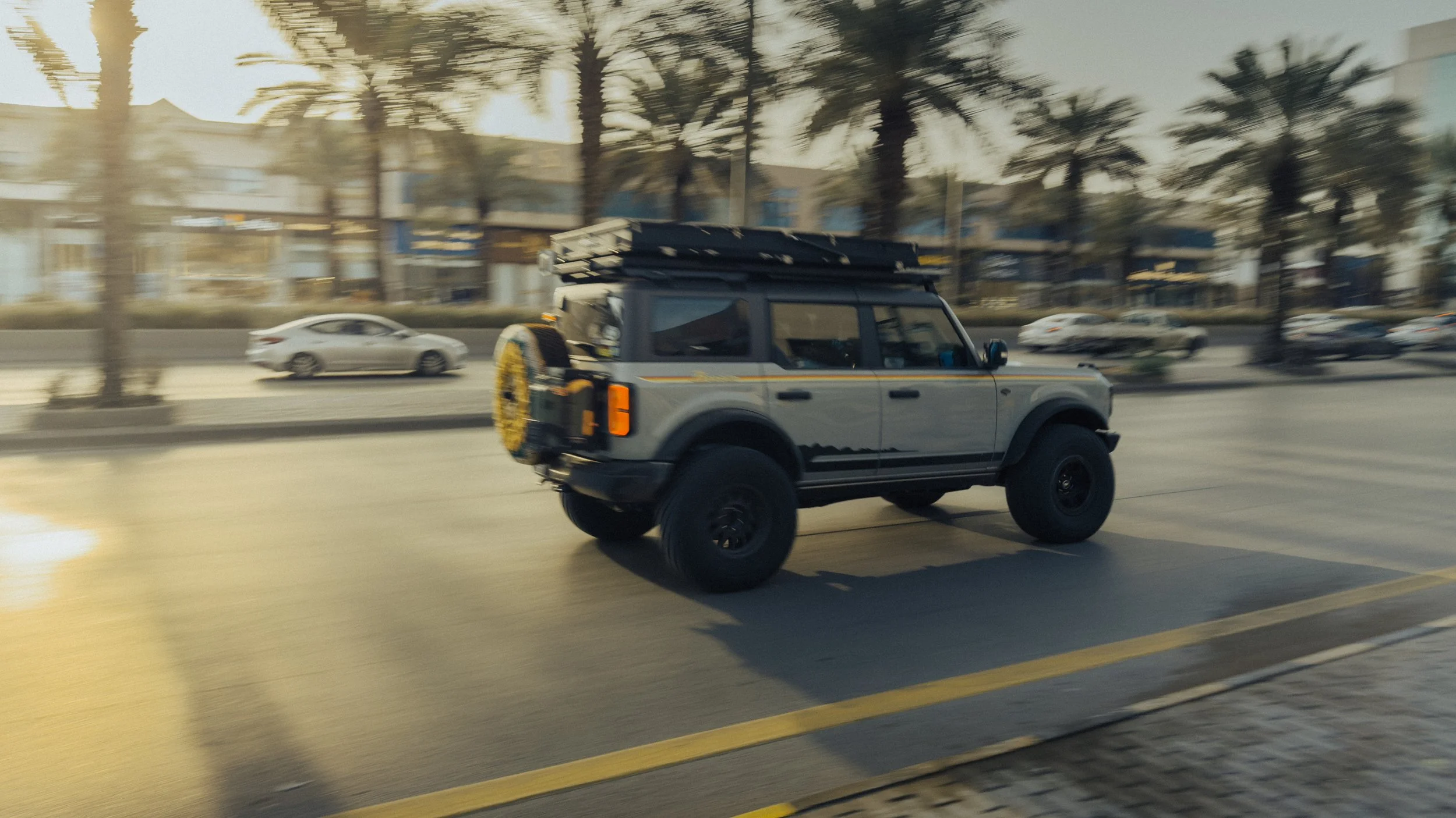 A gray off-road SUV with black wheels driving on a city street during sunset, with palm trees and buildings visible in the background.