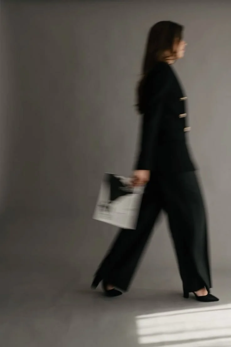 A woman walking in a half profile, dressed in a black suit, holding a magazine or brochure, against a plain gray background.