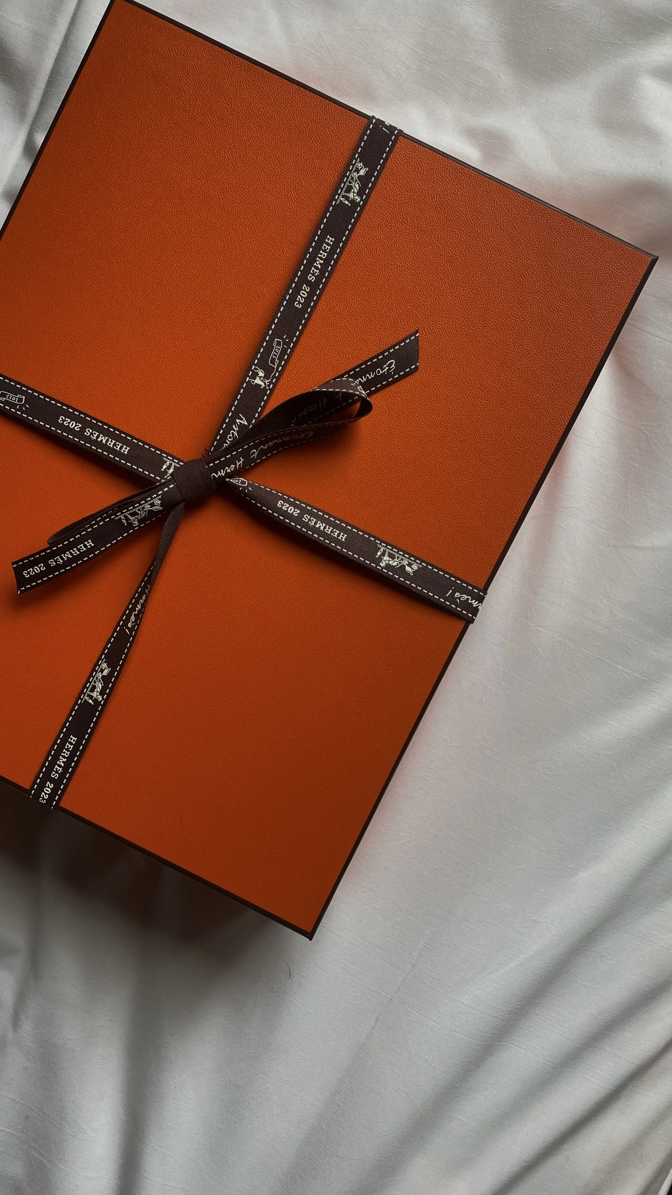 An orange Hermès gift box wrapped with a black ribbon with white text and logos, on a light-colored fabric surface.